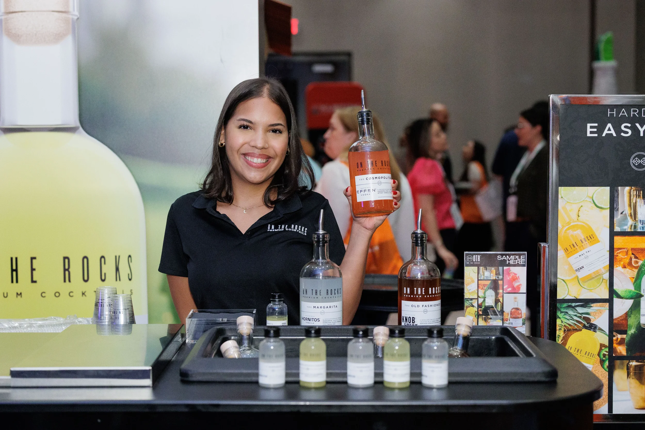 Smiling booth staff holds up branded cocktail bottle at exhibitor table during Orlando corporate trade show