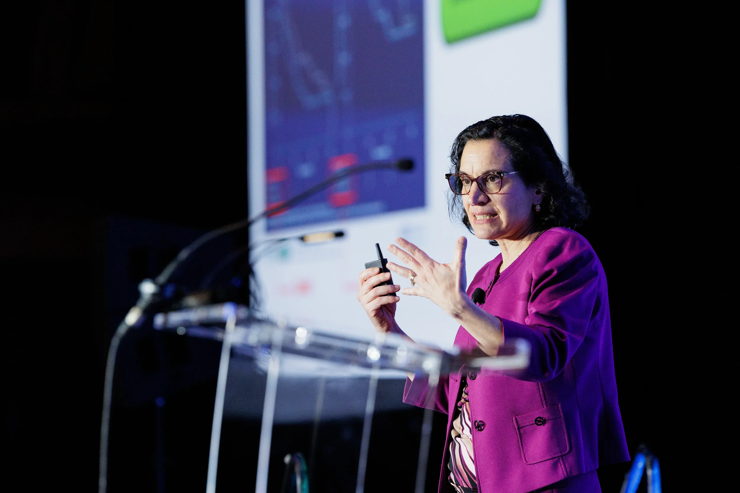 Speaker in magenta jacket gestures with clicker at clear podium during Orlando industry conference