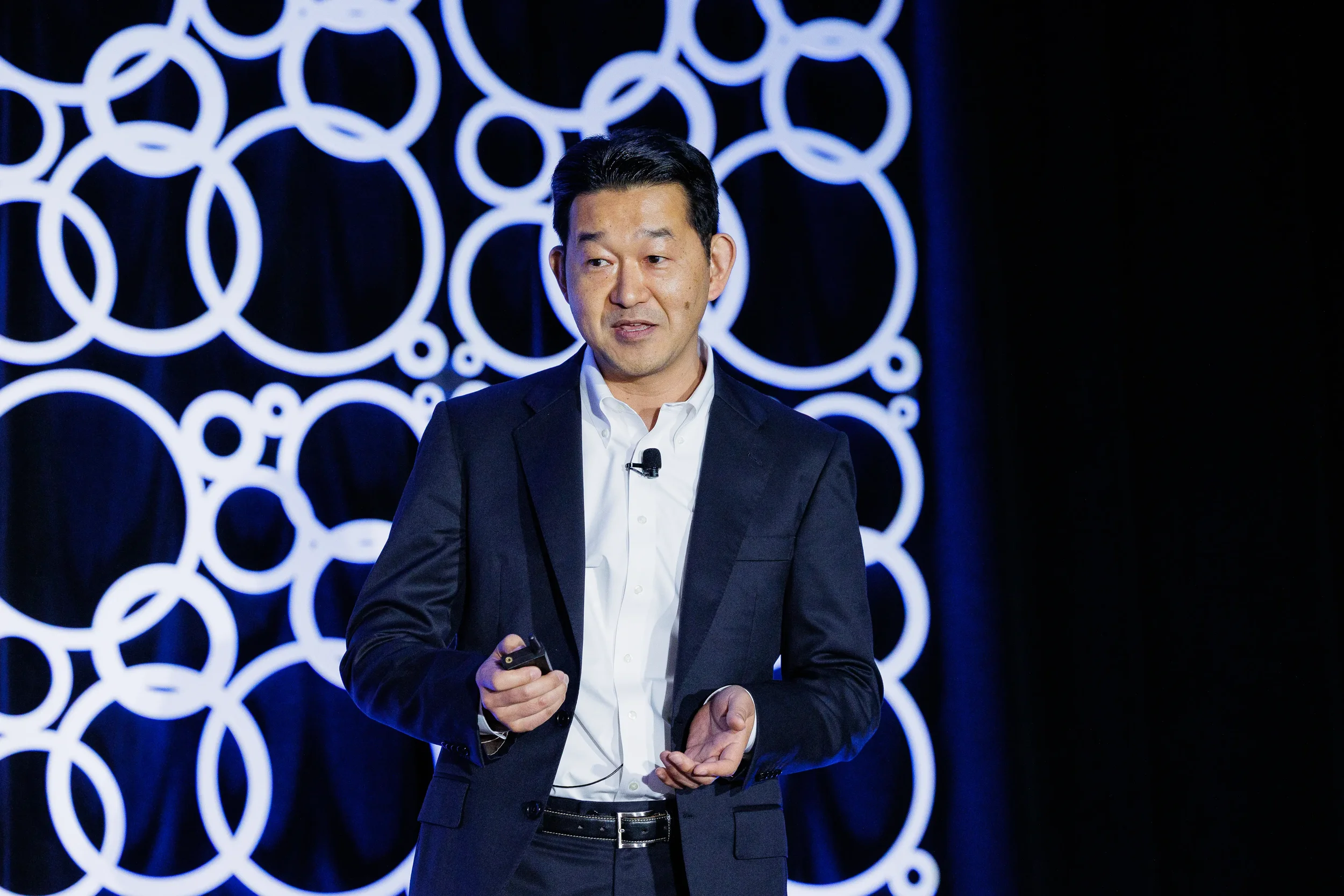 Speaker in navy suit holds slide clicker on stage with circle-patterned backdrop at Chicago conference