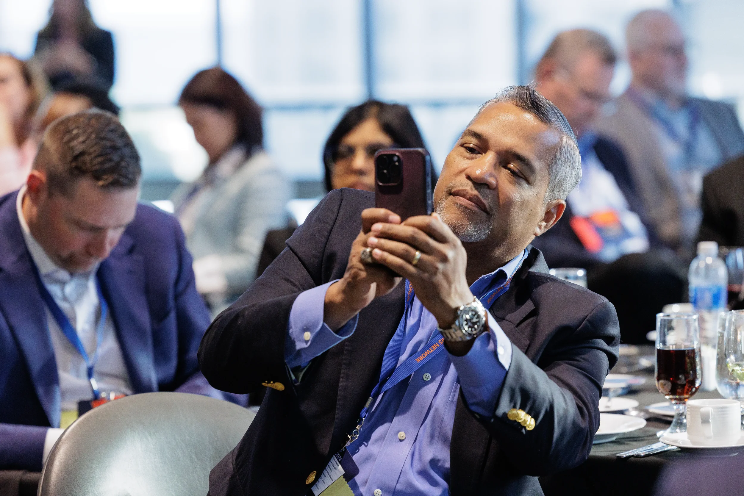 Male attendee with lanyard photographs with phone while seated at Chicago industry conference session