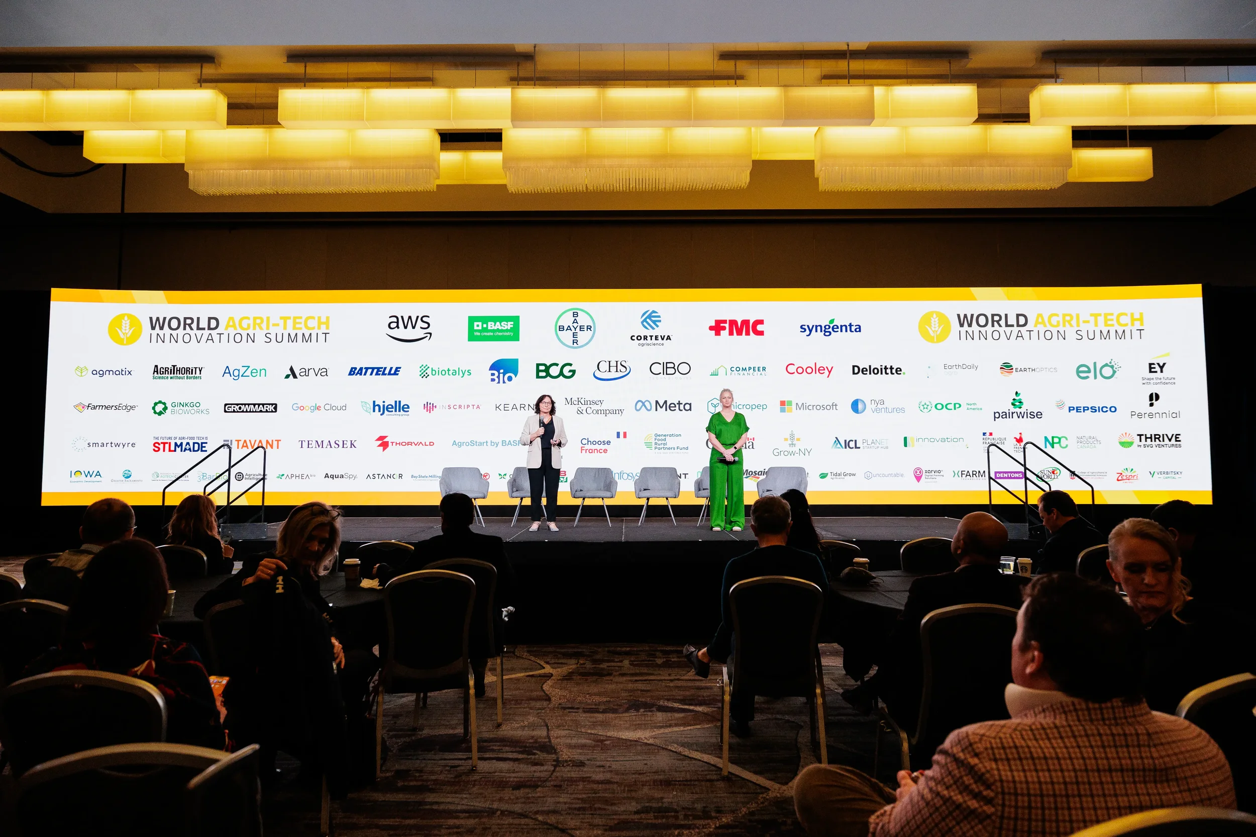 Two speakers present on stage with sponsor logo wall at Orlando industry summit