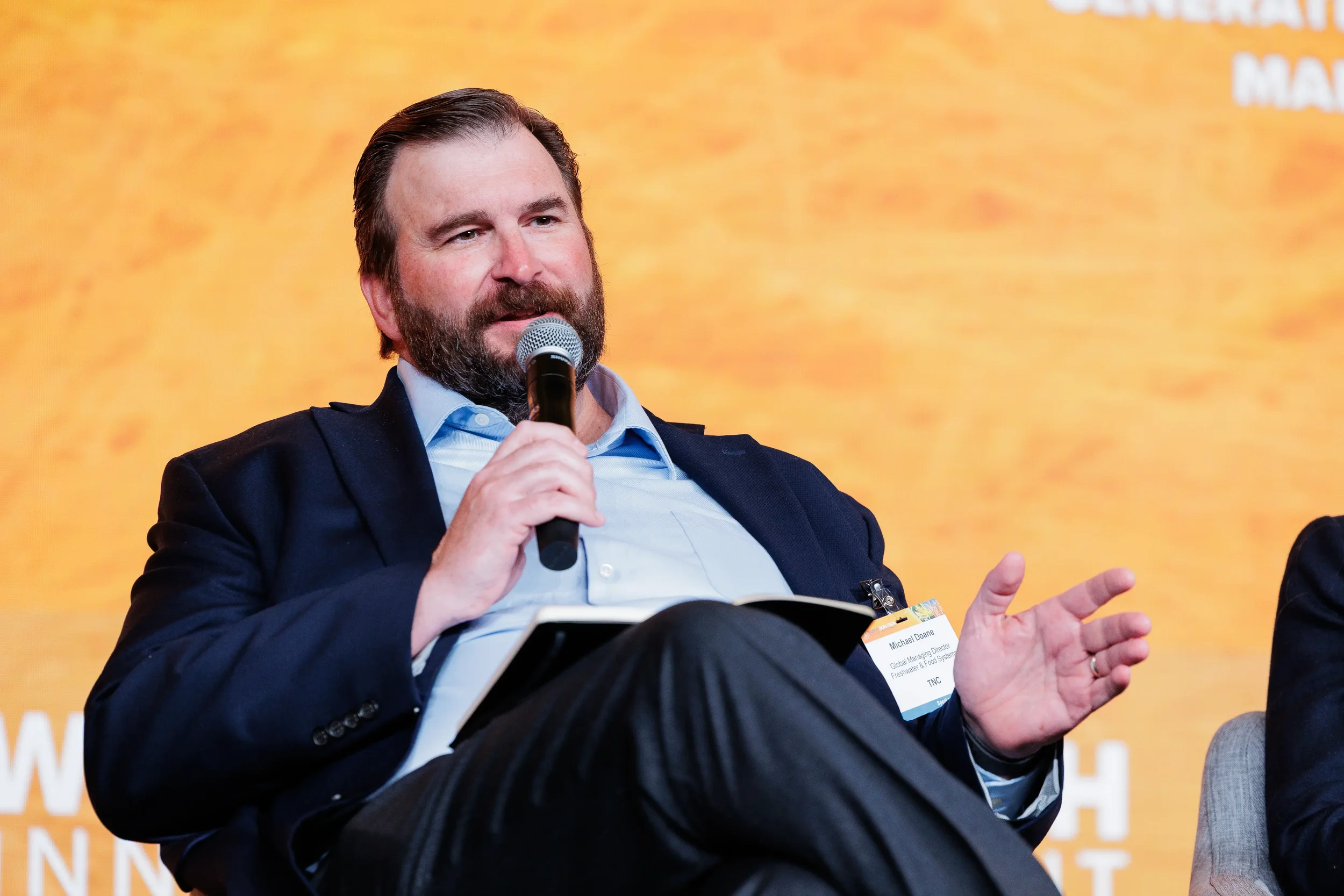 Panelist with name badge speaks into microphone on stage with orange branded backdrop at Chicago conference