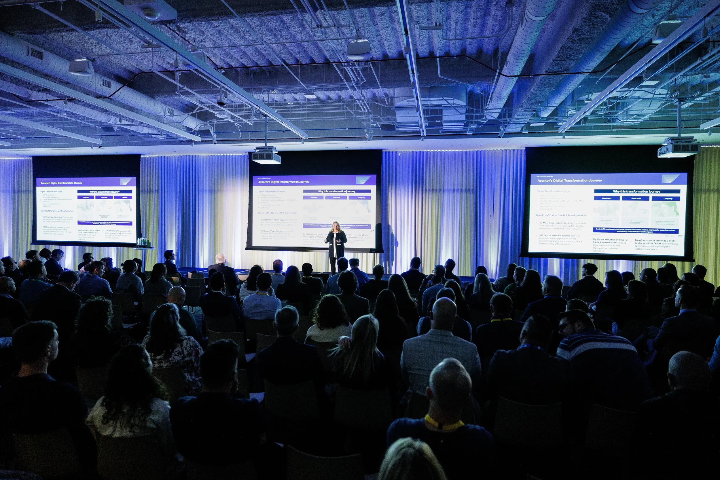 Presenter speaks on stage before three screens and packed audience at Orlando industry conference