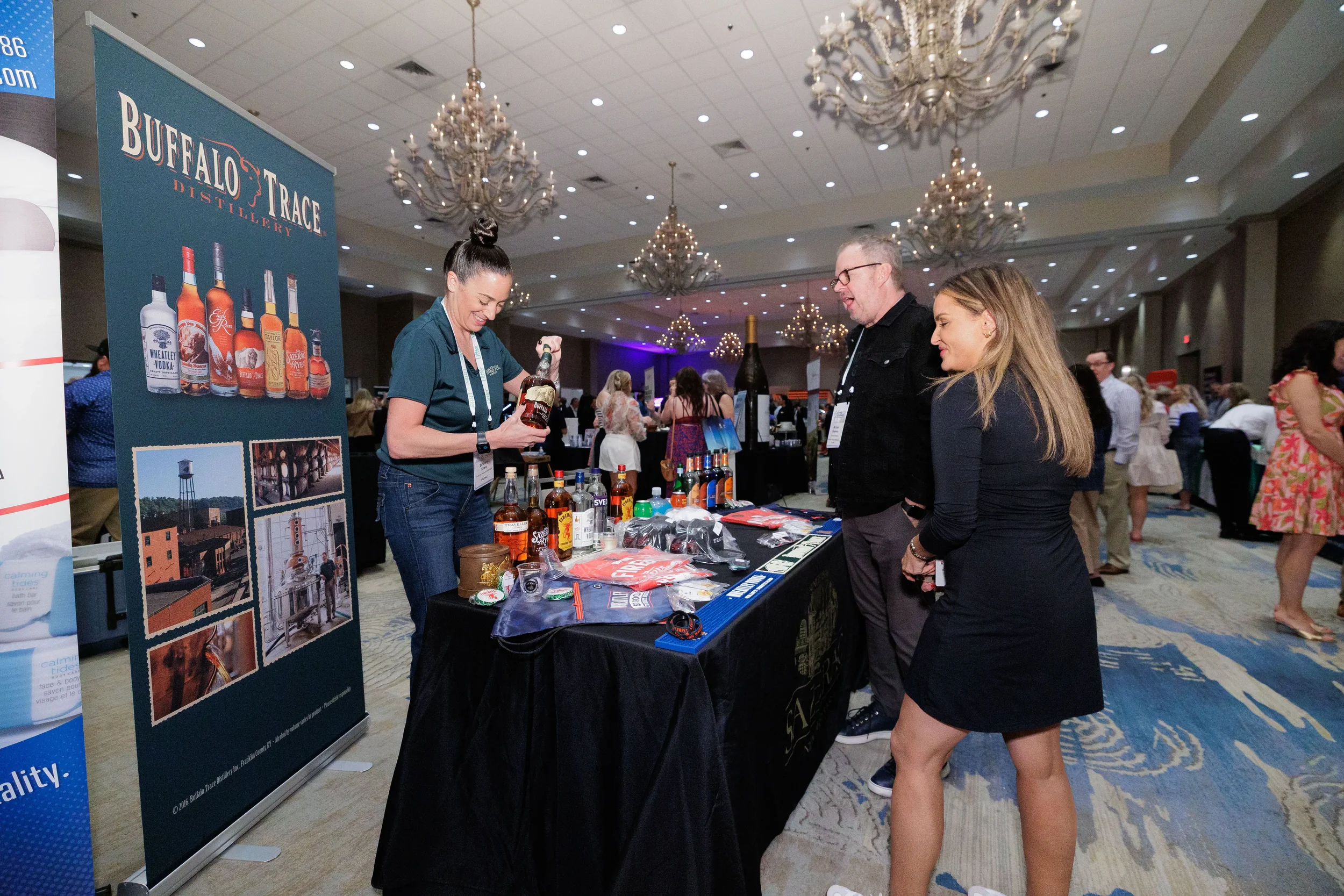 Booth staff pours drinks for two attendees at exhibitor table in chandelier ballroom at Orlando trade show