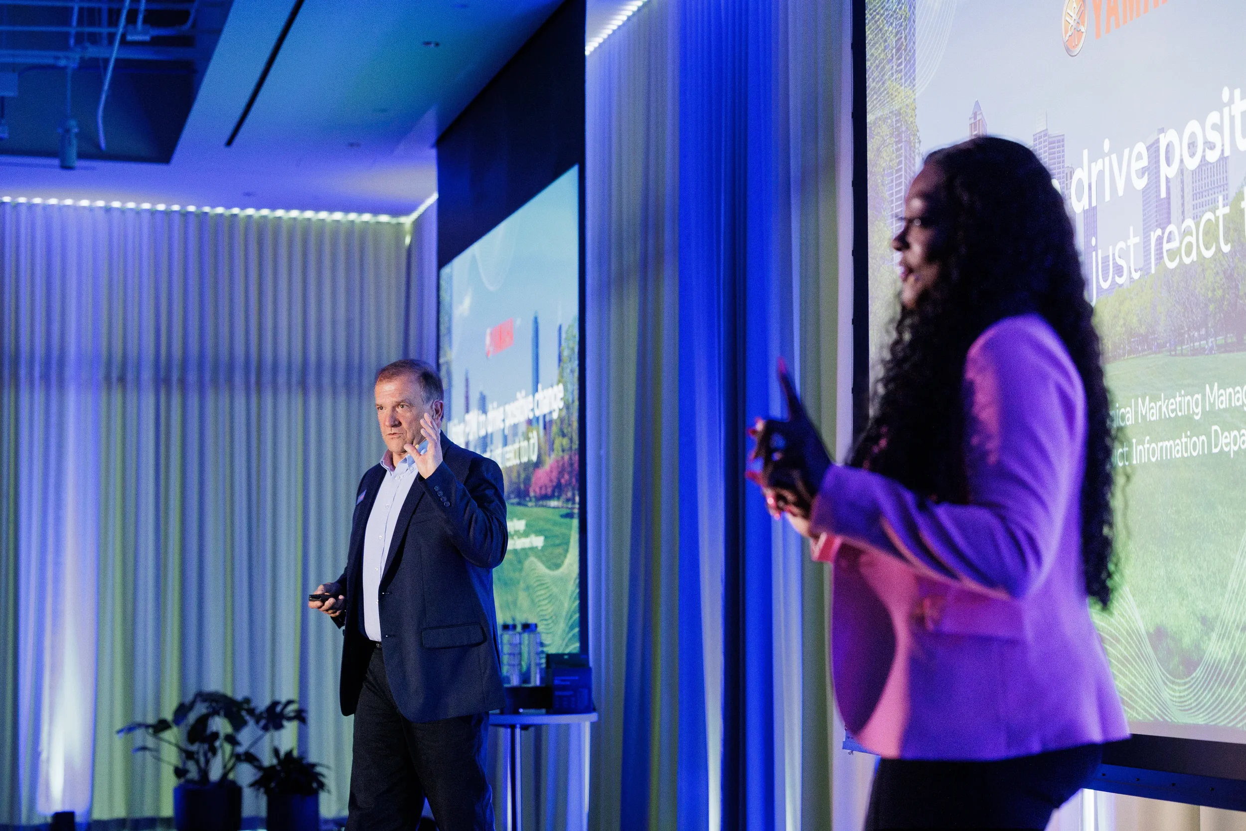 Two speakers present together on stage with blue curtain lighting at Orlando corporate conference
