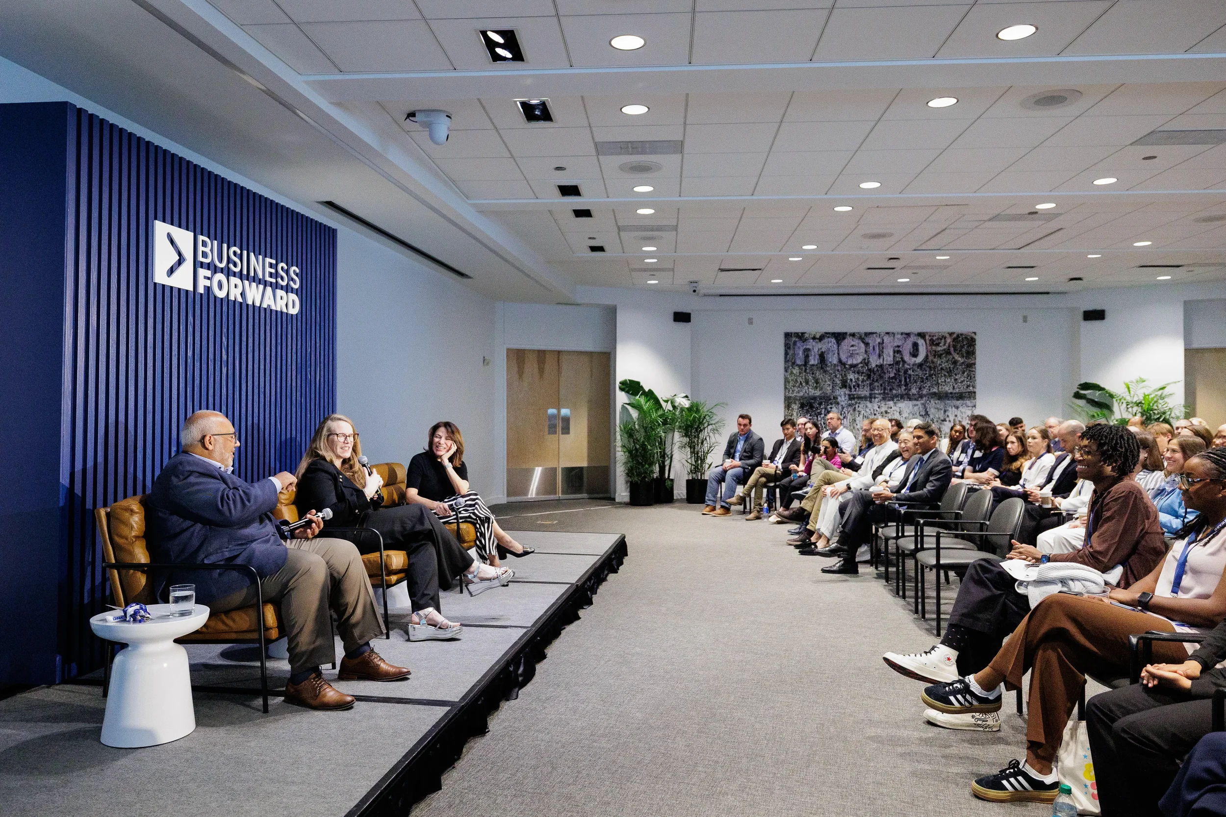 Three panelists on low stage face seated audience rows beside Business Forward branded wall at Chicago corporate conference venue
