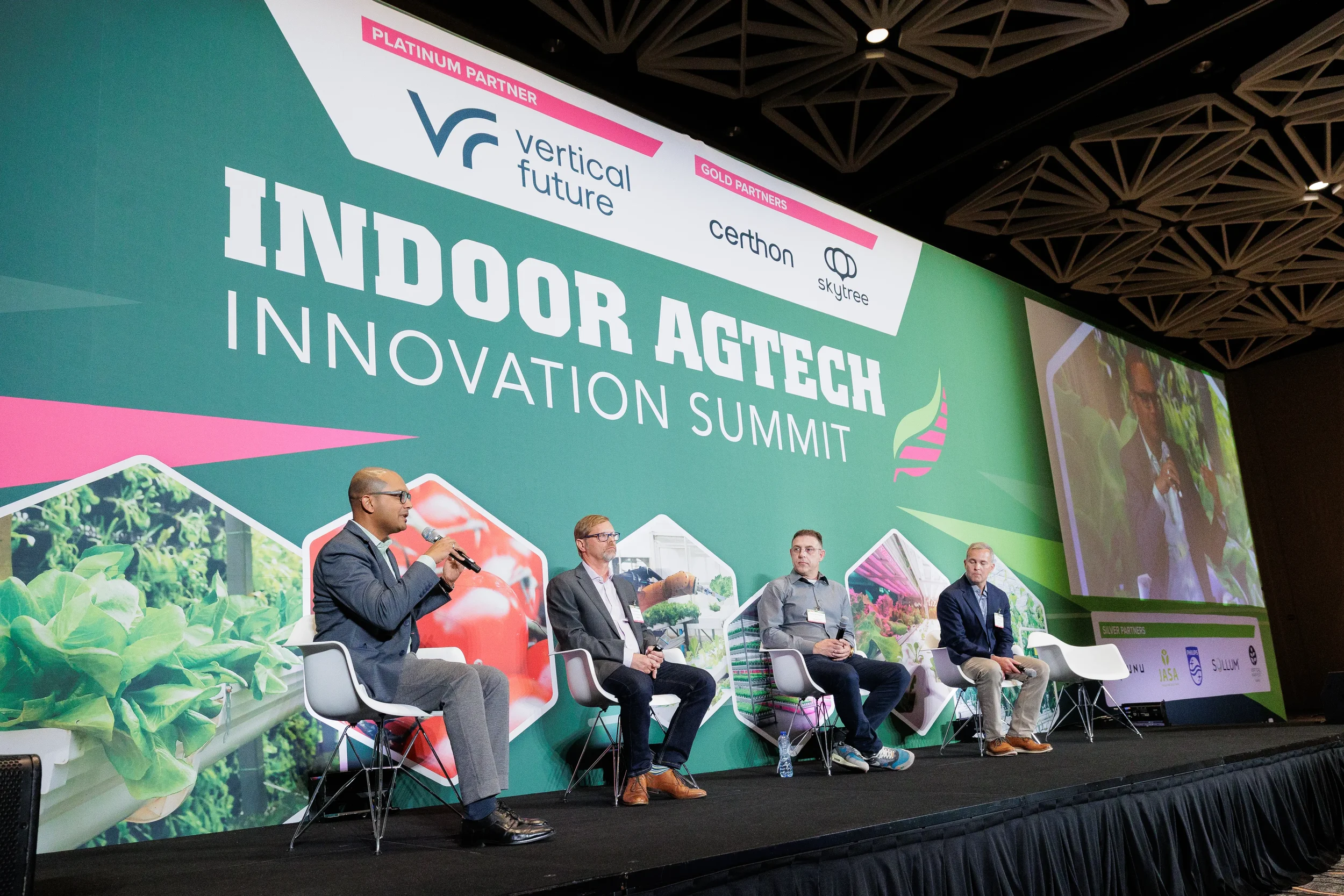 Panelist speaks into microphone while three fellow speakers listen on stage in front of large branded backdrop at Orlando industry conference