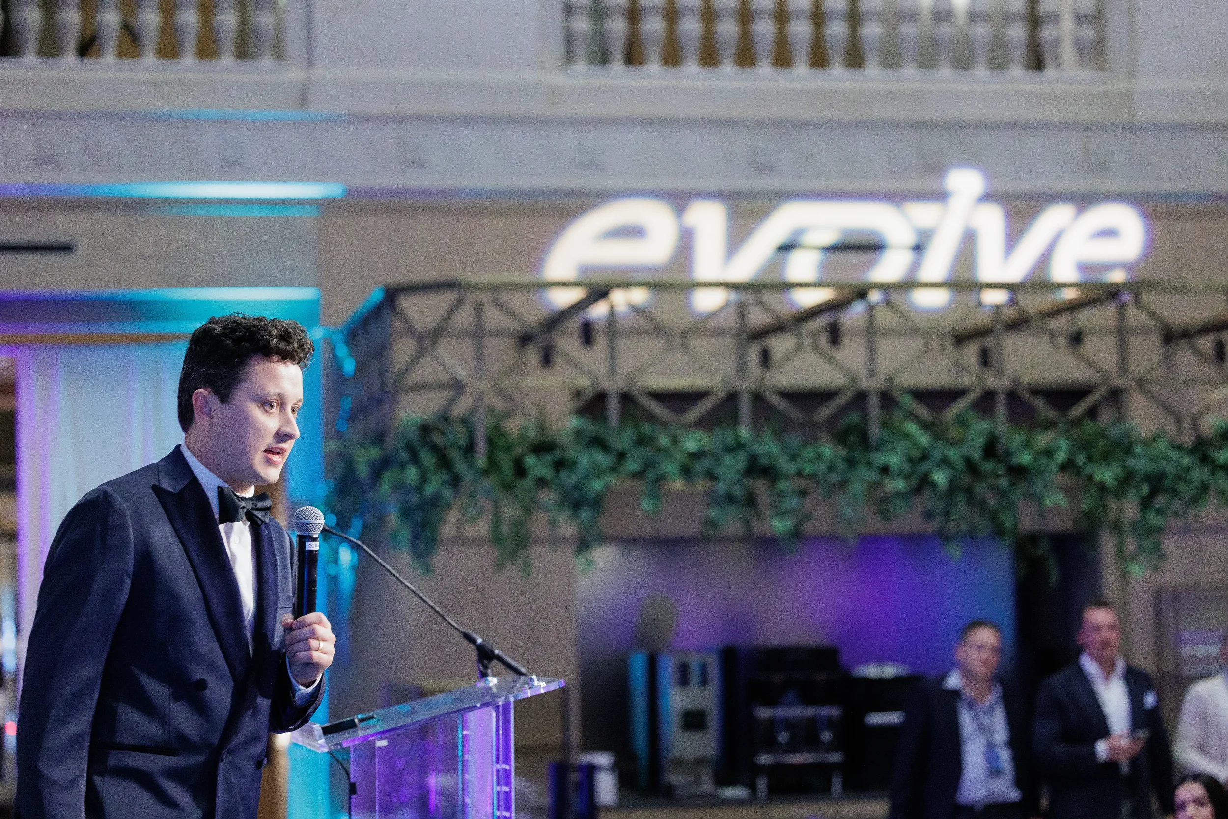 Emcee in tuxedo speaks at podium with glowing brand sign in background at Orlando corporate event