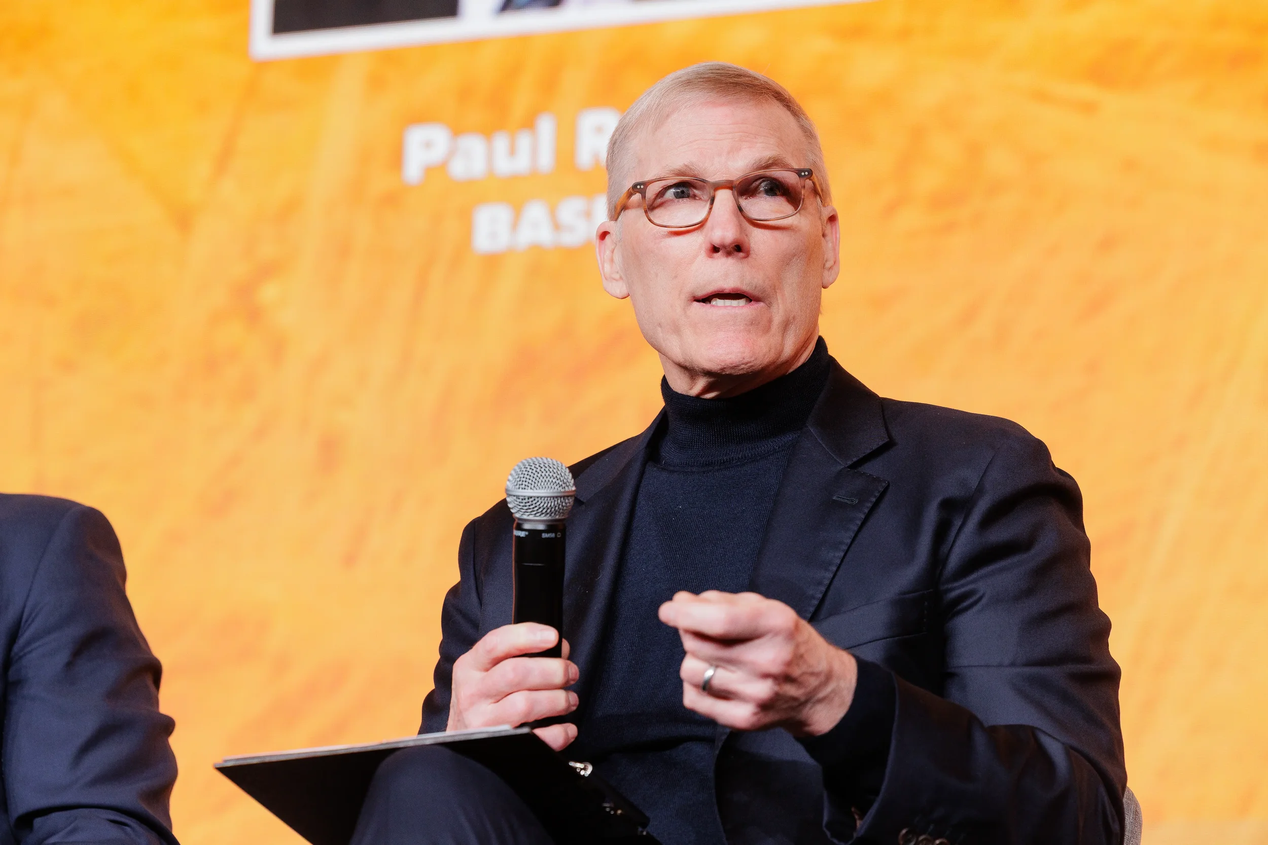 Male panelist in turtleneck and glasses holds microphone and gestures on stage at Orlando industry conference