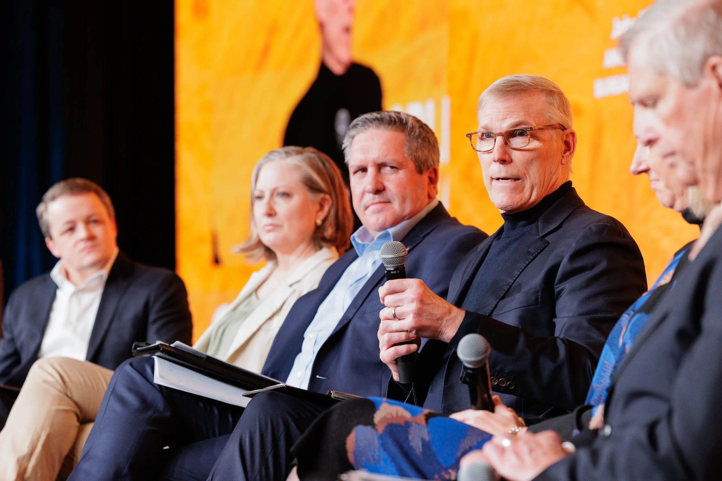 Panelist speaks into microphone seated in row on stage with orange branded backdrop at Orlando conference