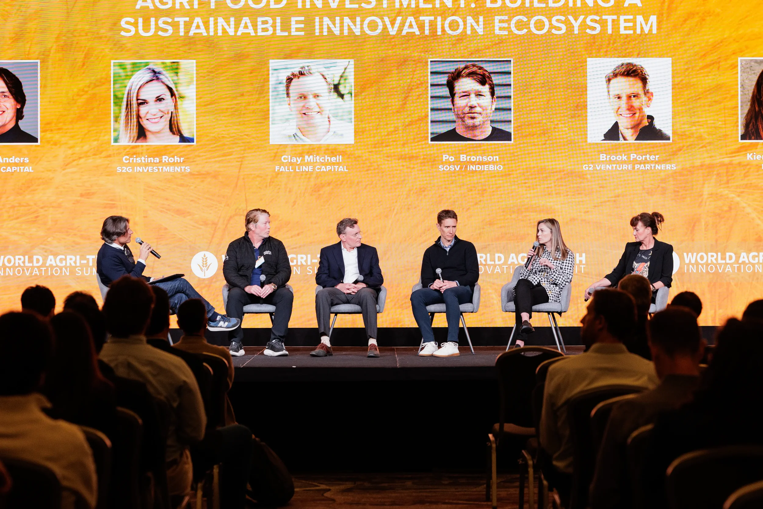 Large panel of six speakers seated on branded stage with audience silhouettes at Orlando corporate conference