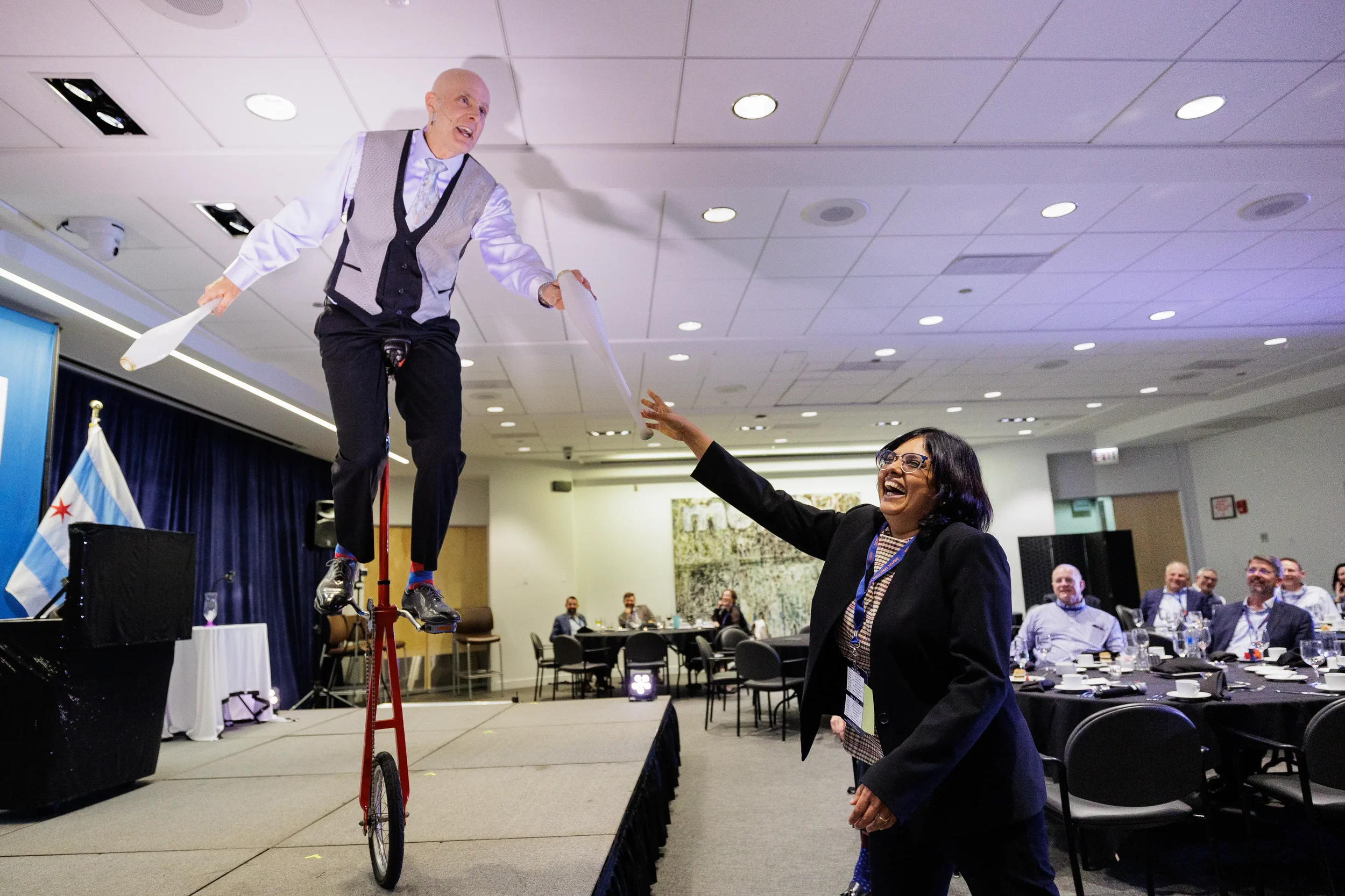 Entertainer rides unicycle while juggling clubs with laughing attendee on stage at Orlando corporate conference