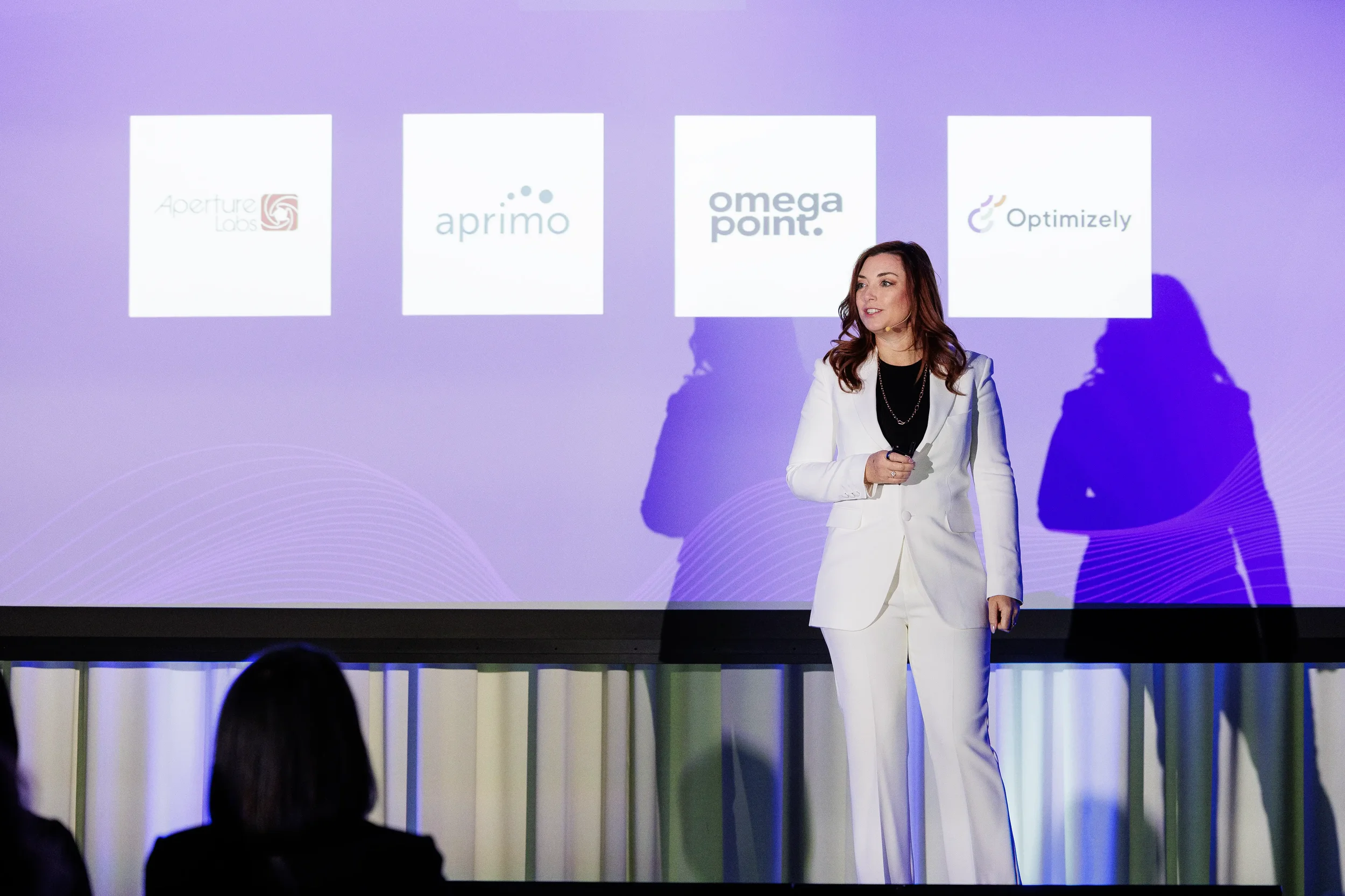 Speaker in white suit presents on stage with partner logos on screen at Orlando corporate conference