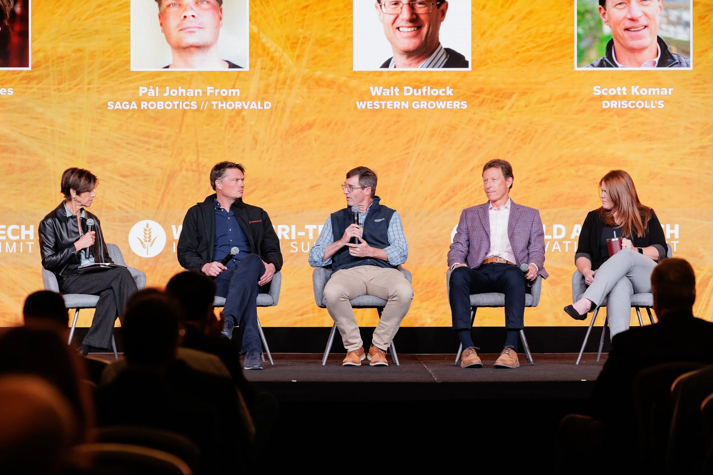 Five panelists seated on branded orange stage with audience silhouette in foreground at Orlando industry conference