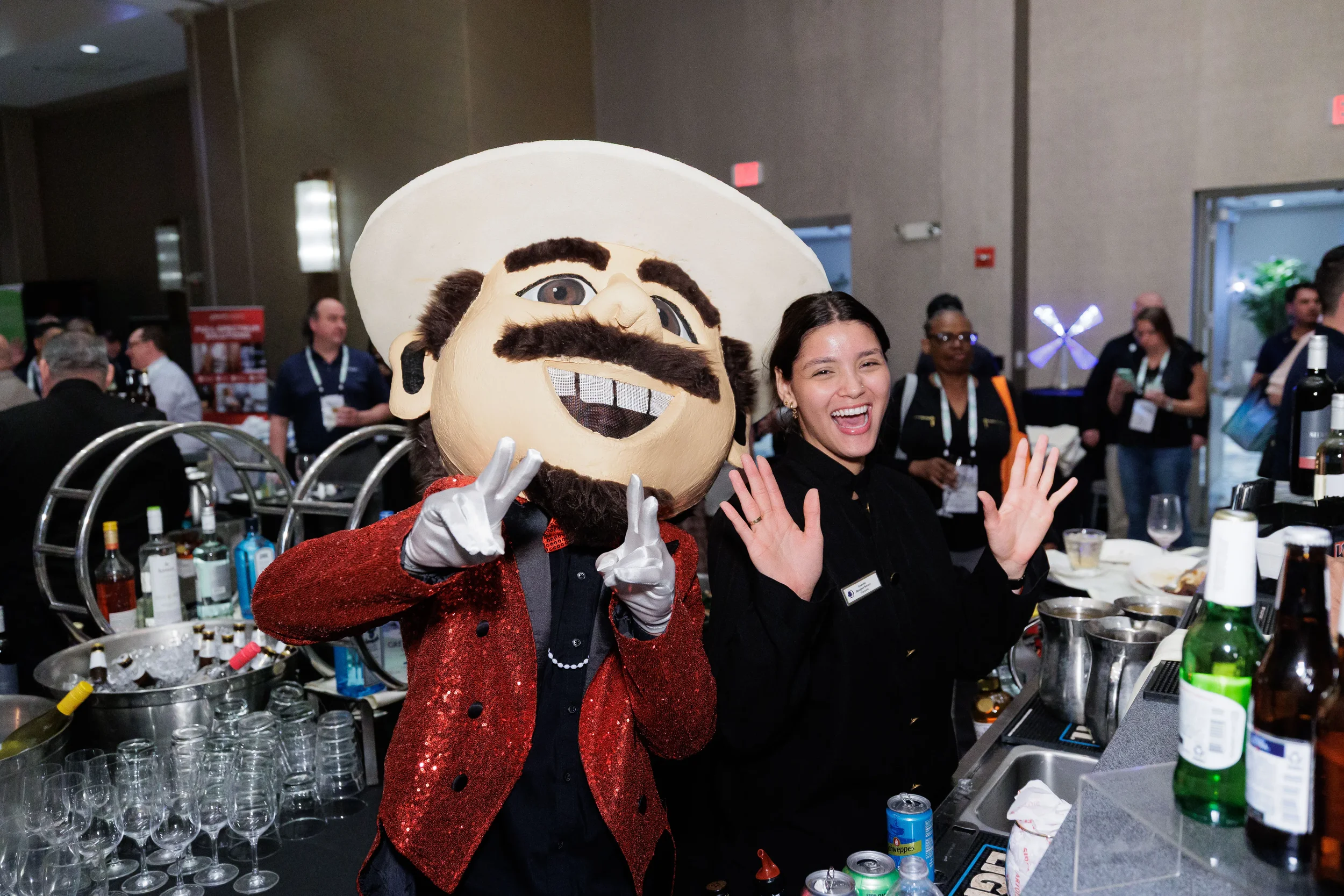 Oversized mascot character poses with laughing bartender at bar during Orlando corporate conference expo