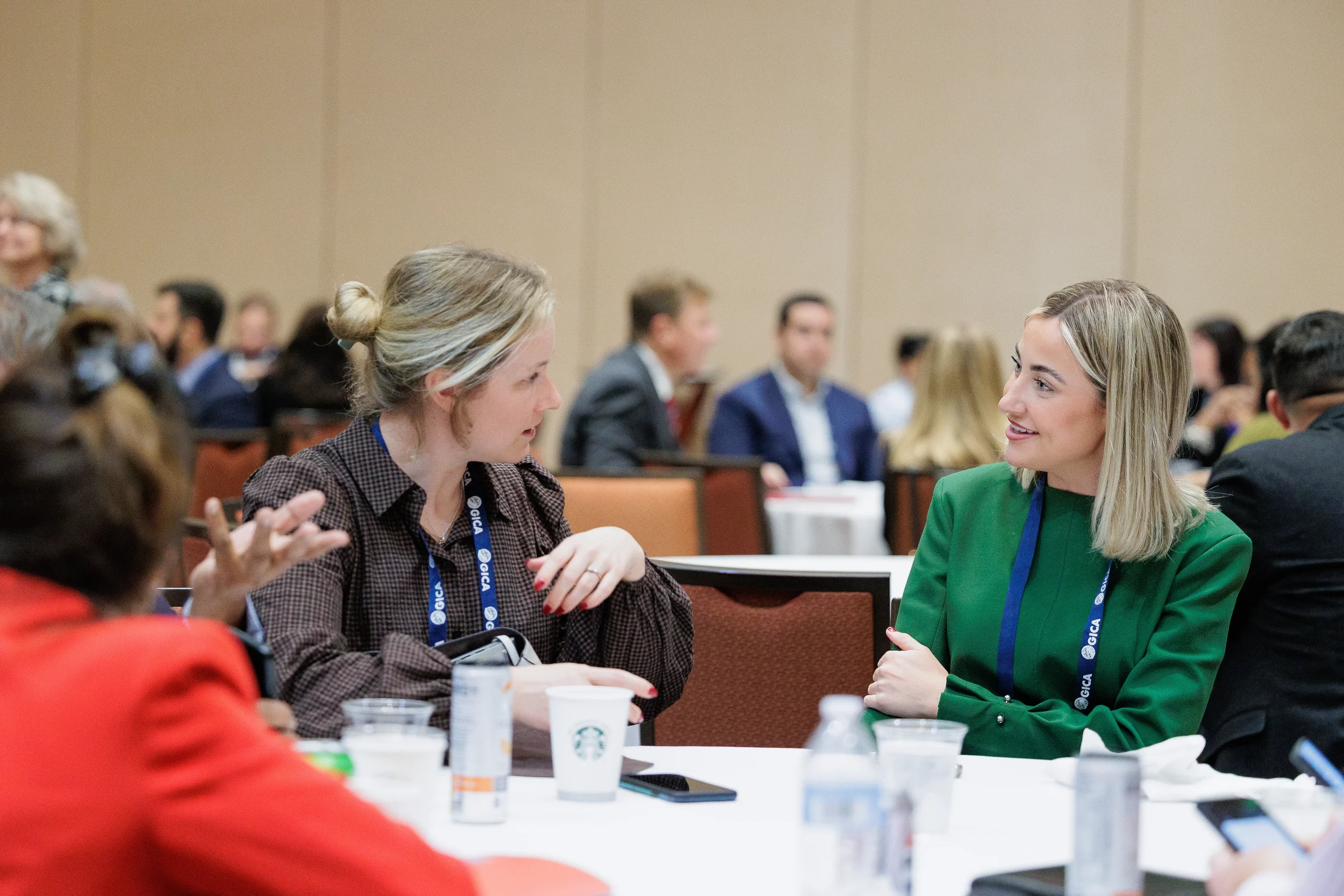 Two women in professional attire speak at a conference table. Blurred attendees sit behind them in an Orlando conference room