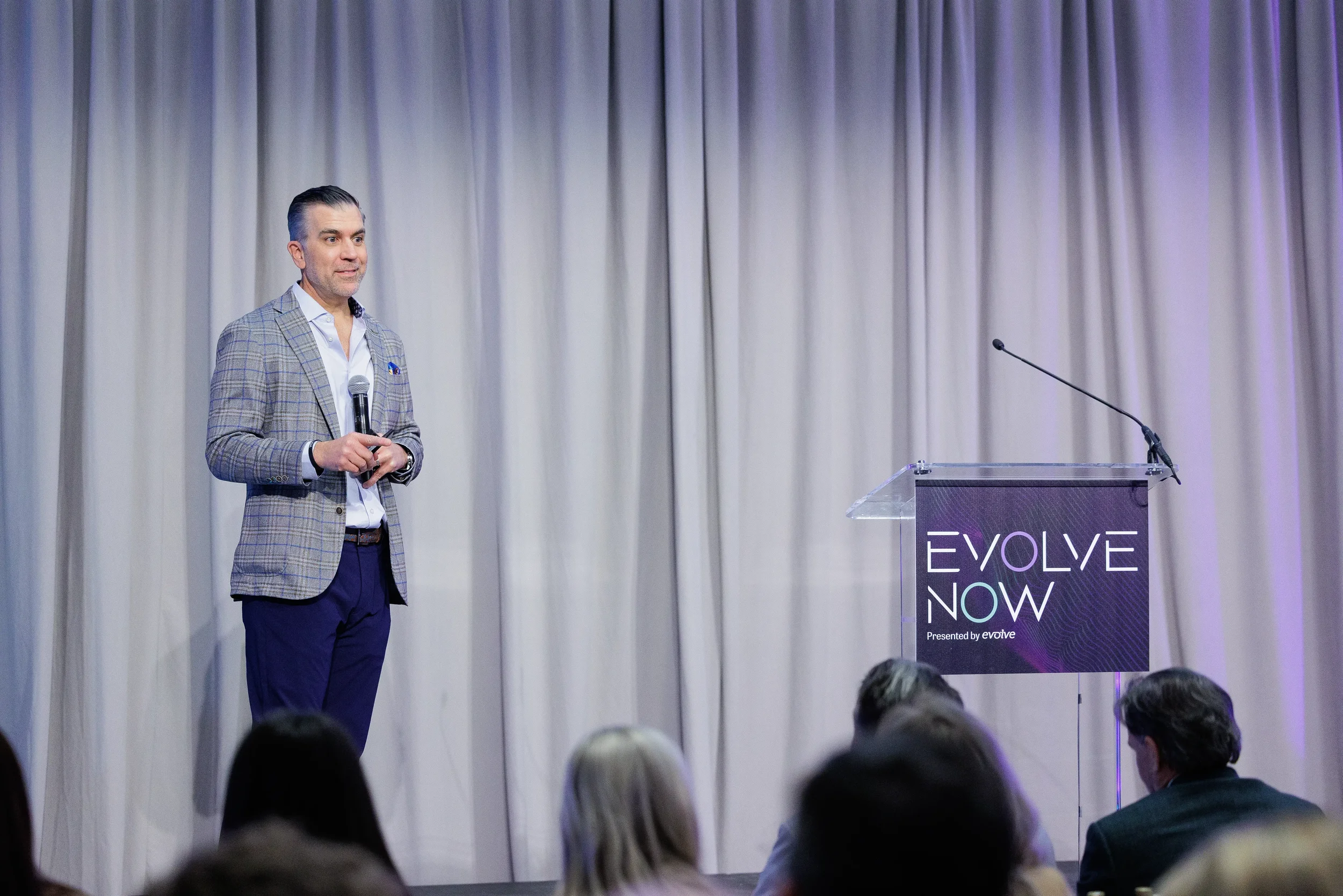 Speaker in plaid blazer presents on stage beside branded podium with audience in foreground at Chicago conference