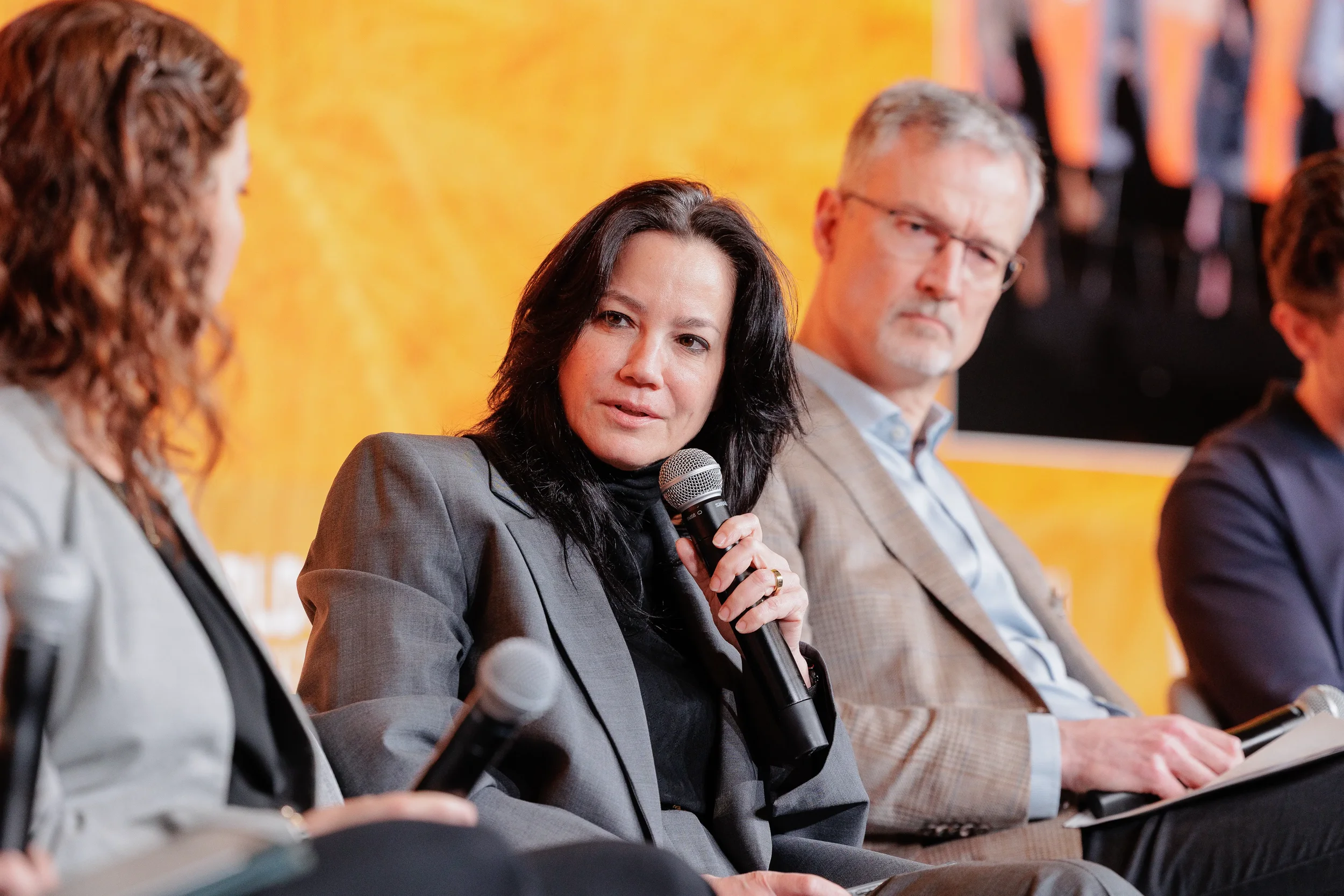 Female panelist in gray blazer holds microphone on stage before orange backdrop at Orlando corporate conference