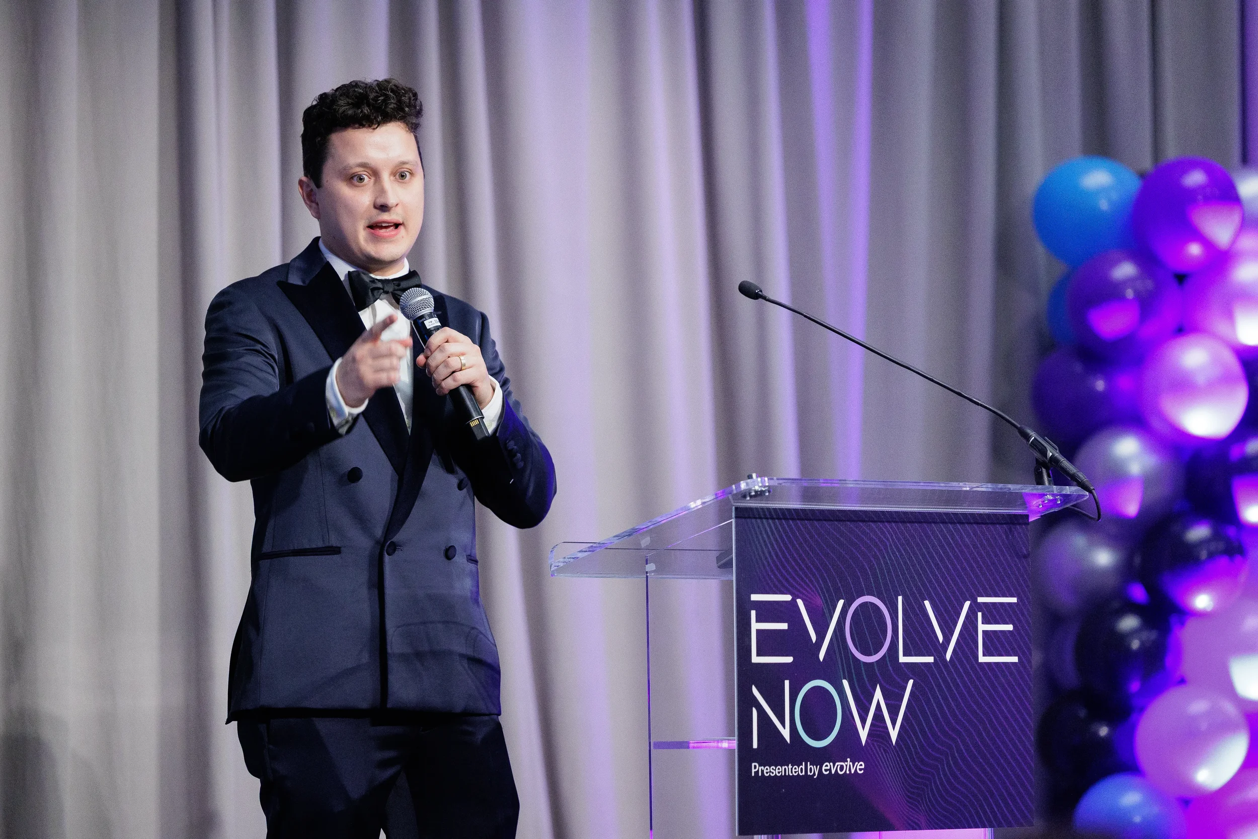 Speaker in tuxedo points to audience while presenting at branded podium during Orlando corporate event
