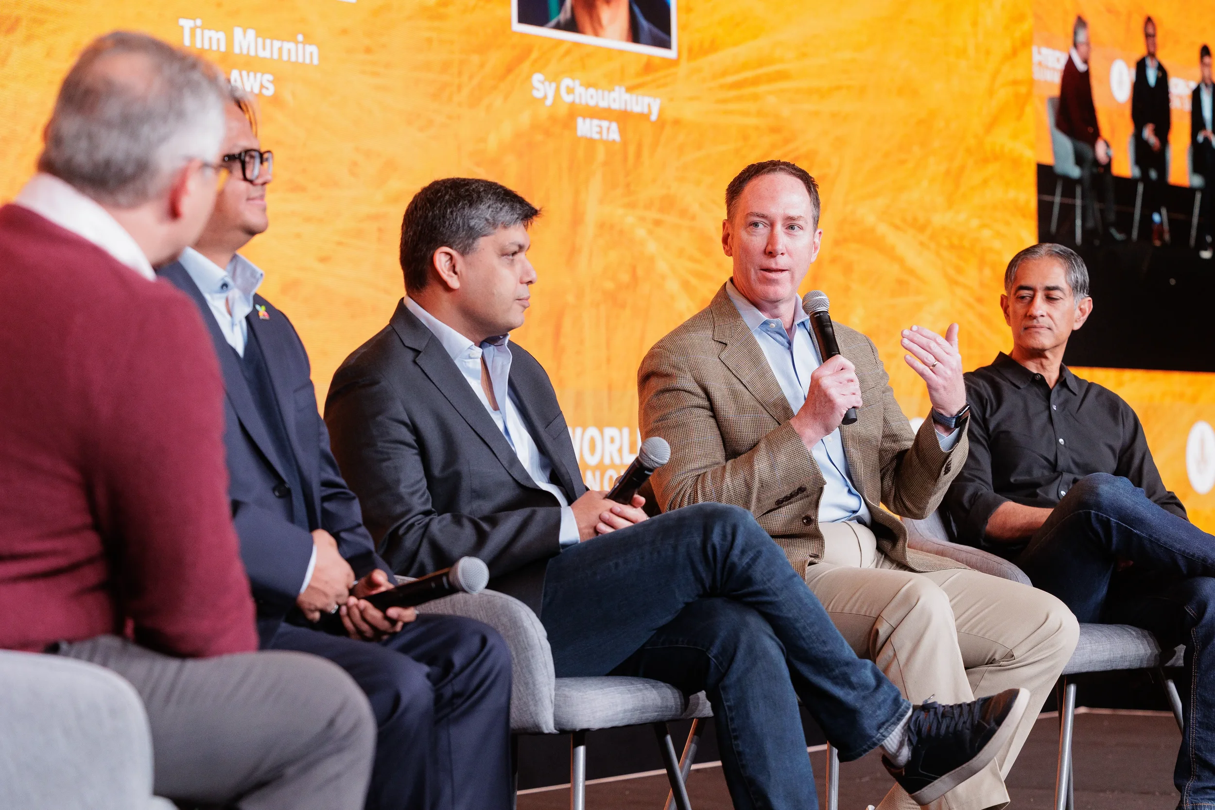 Five panelists seated on stage with microphones in front of orange branded backdrop at Chicago corporate conference