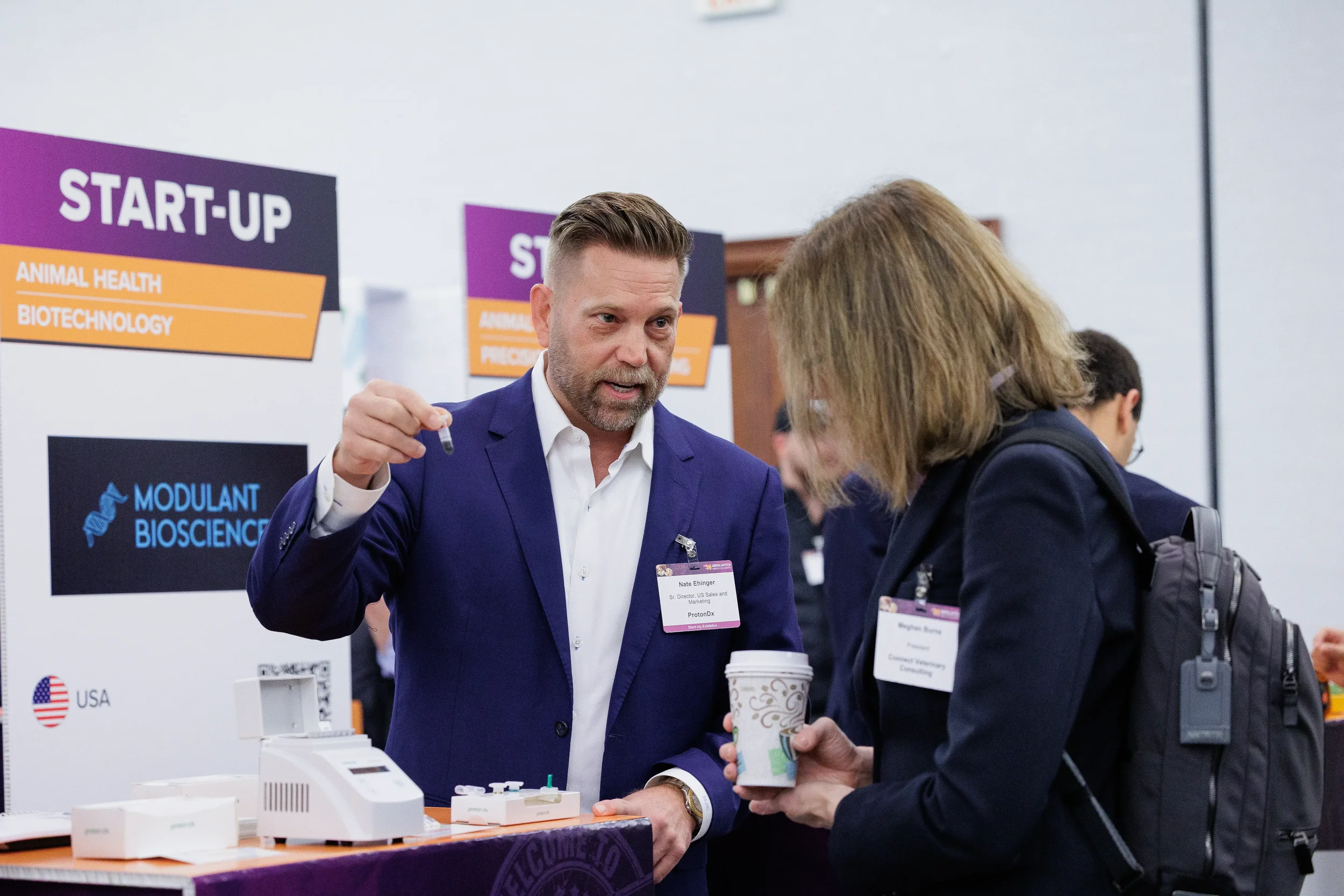 Attendee demonstrates biotech product to visitor at startup booth during Orlando trade show