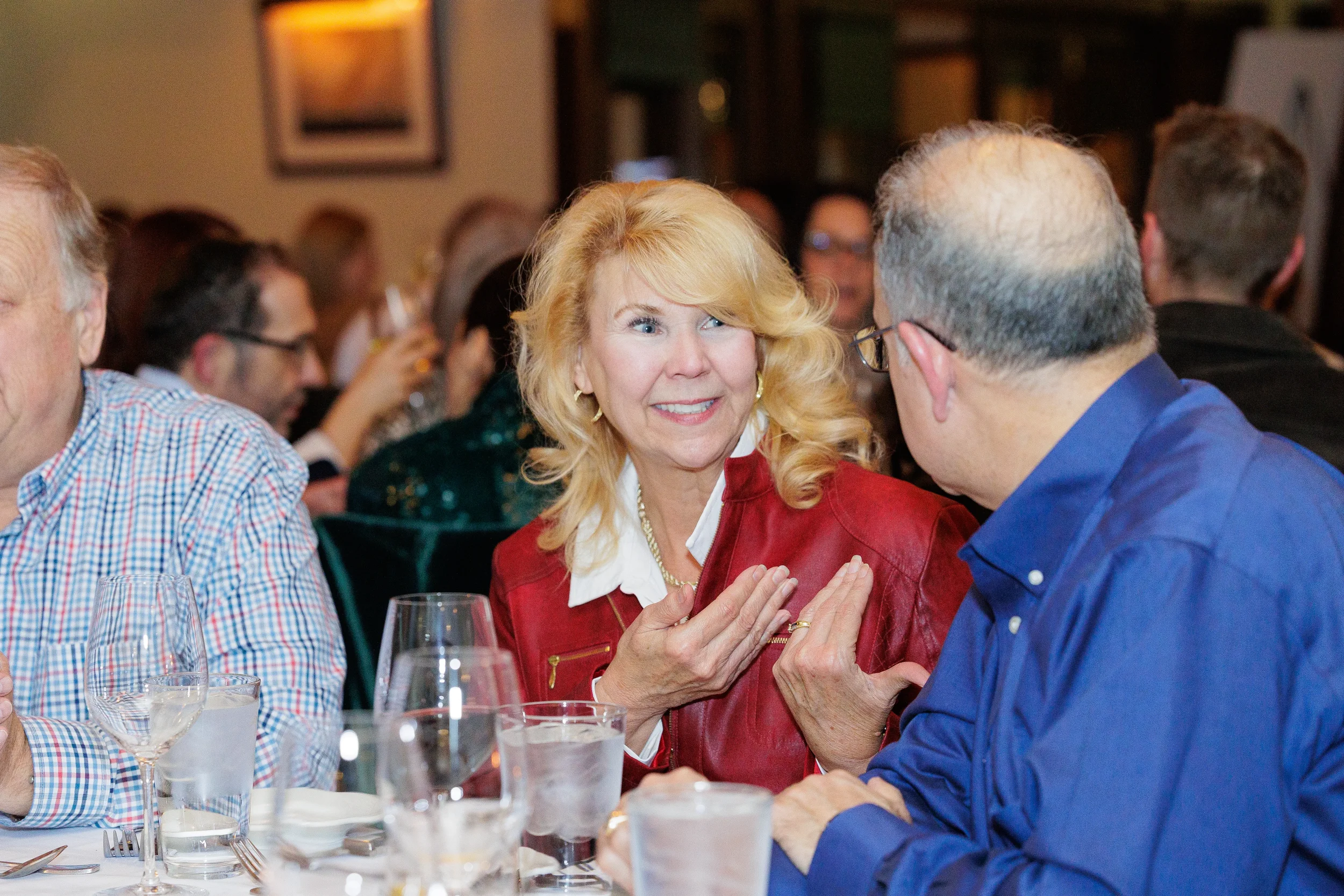 A blond woman in a red jacket smiles and gestures while seated at a dining table across a man in a blue shirt in Orlando