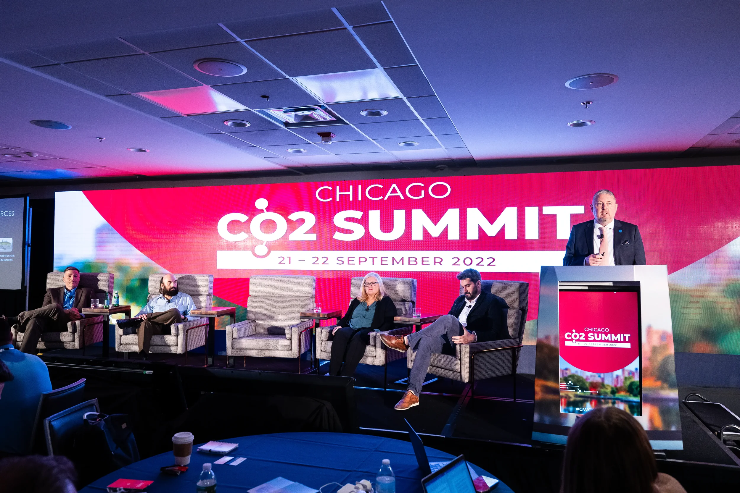 Four panelists and moderator at podium on branded stage during Chicago industry summit panel discussion