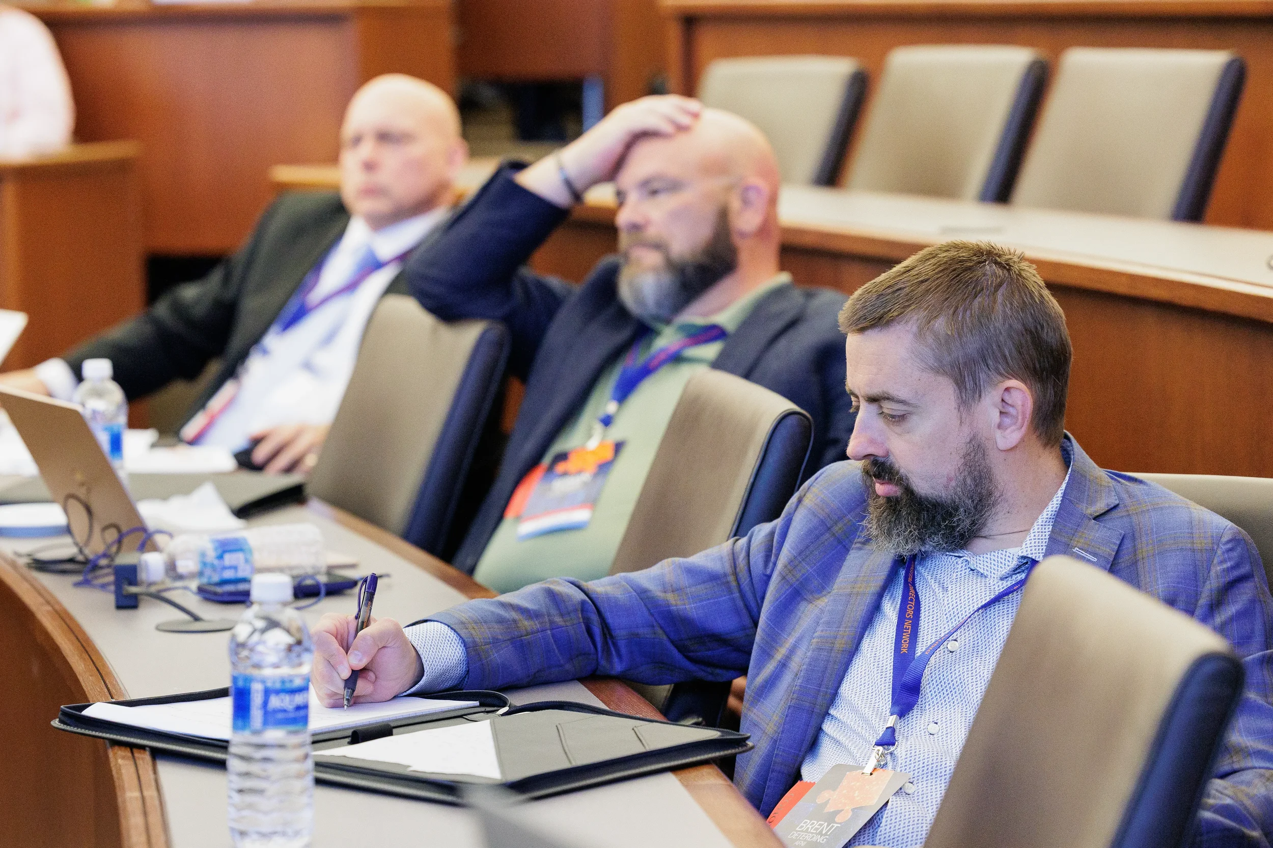 Three male attendees with lanyards take notes and listen in tiered room at Orlando industry conference