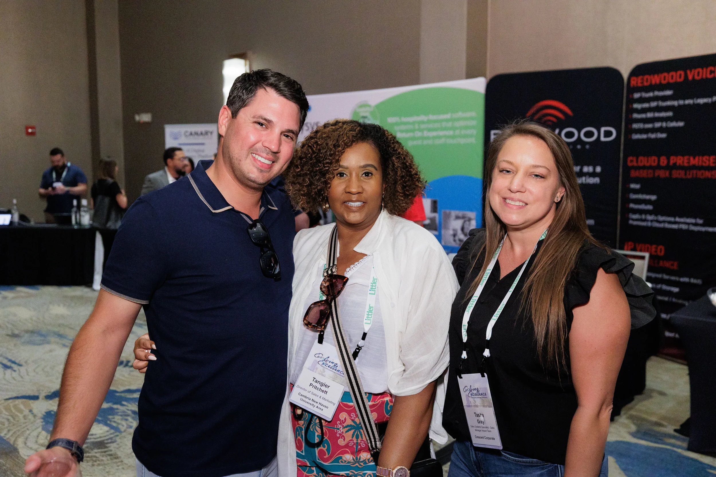 Three attendees with lanyards pose together near sponsor booths at Orlando industry conference trade show