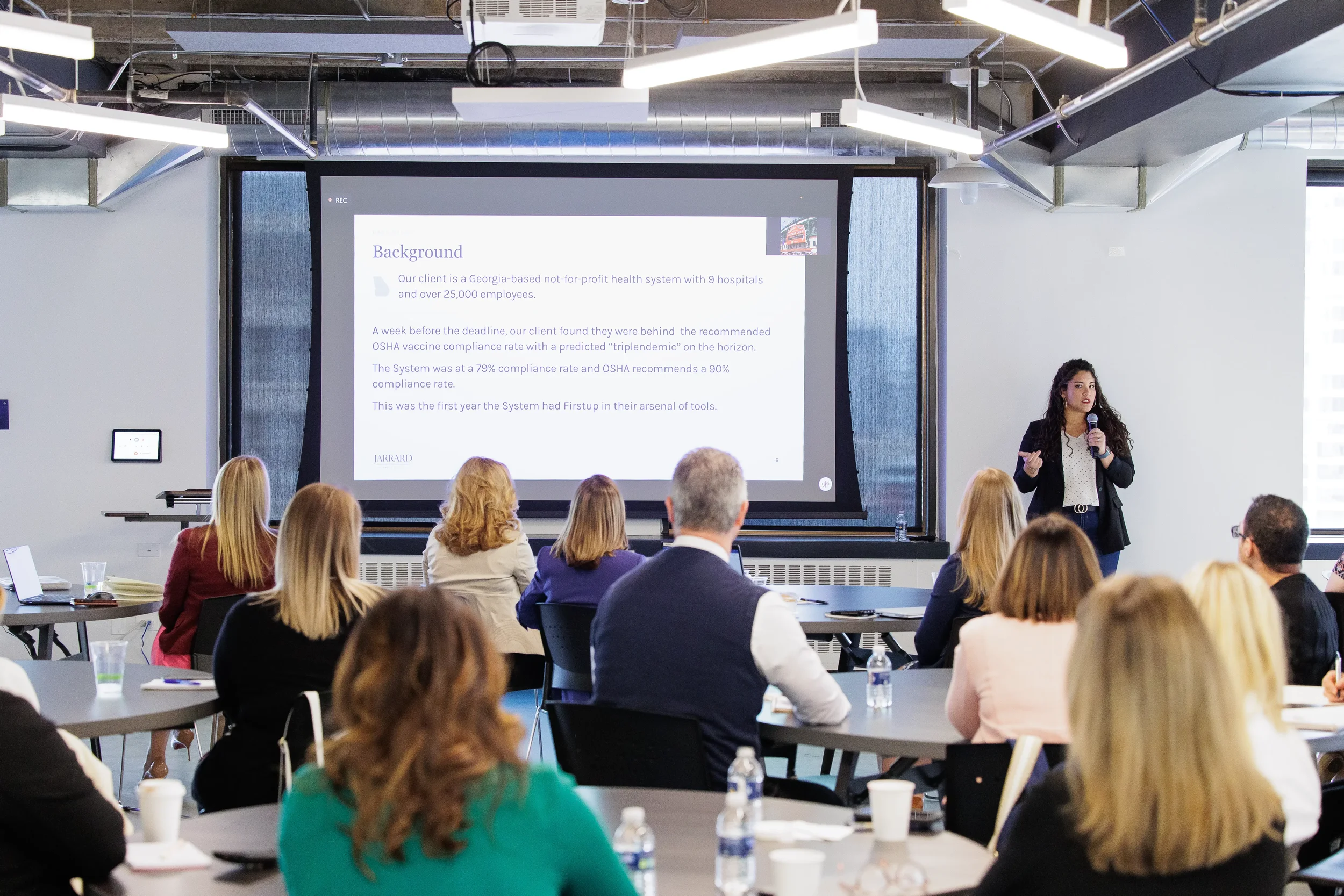 Female speaker presents to a full room of seated attendees beside large projection screen at Orlando conference