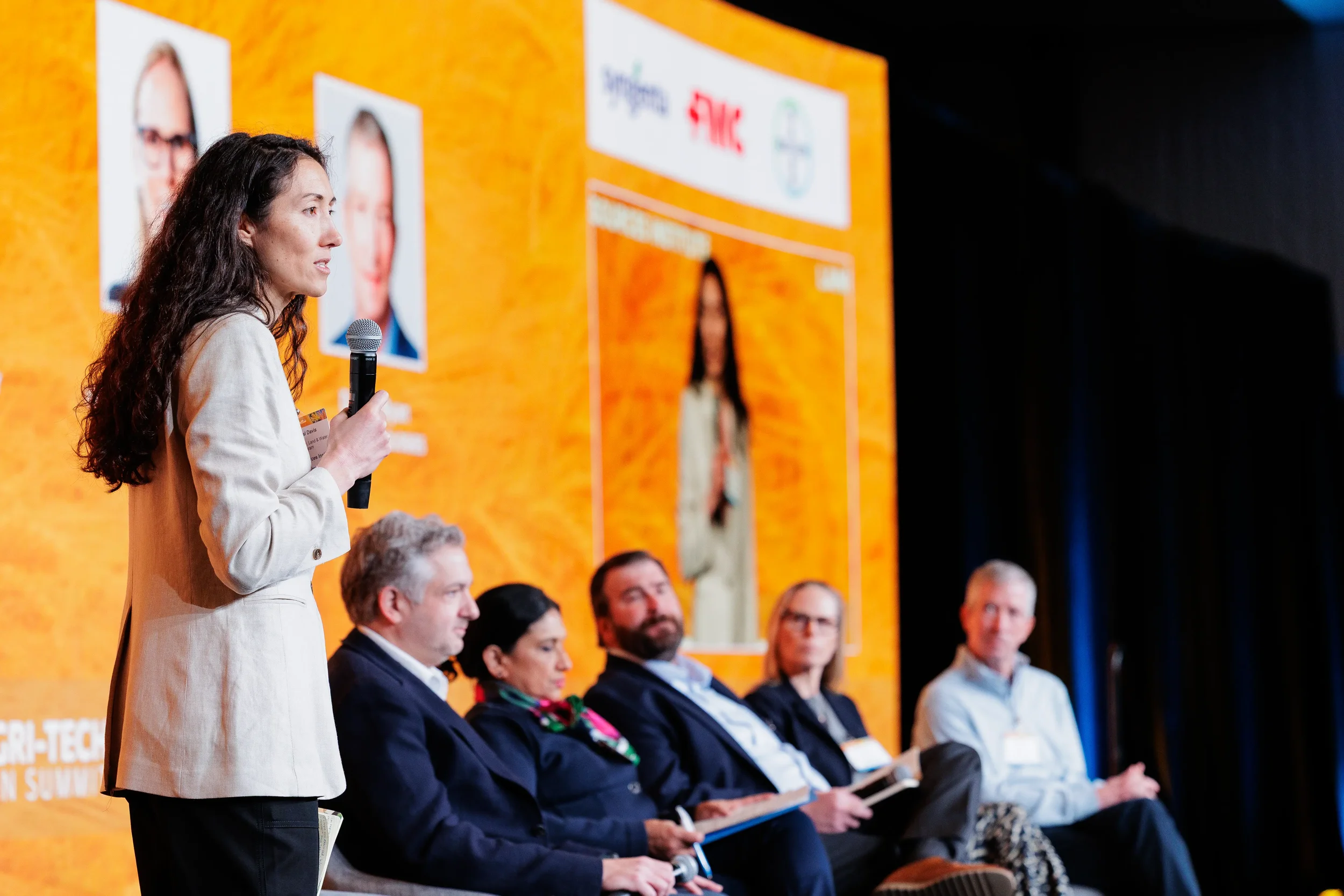Standing moderator holds microphone before seated panel on branded orange stage at Orlando industry conference