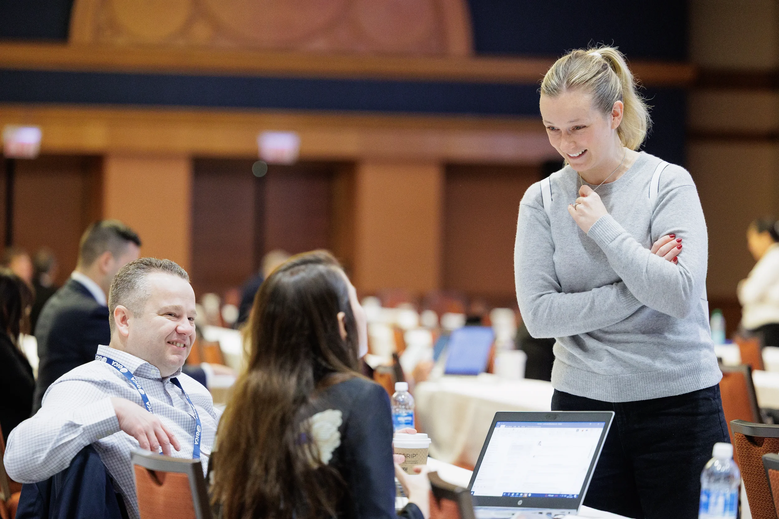 Three professionals talk at tables in a large Orlando conference hall with a laptop visible in the foreground.