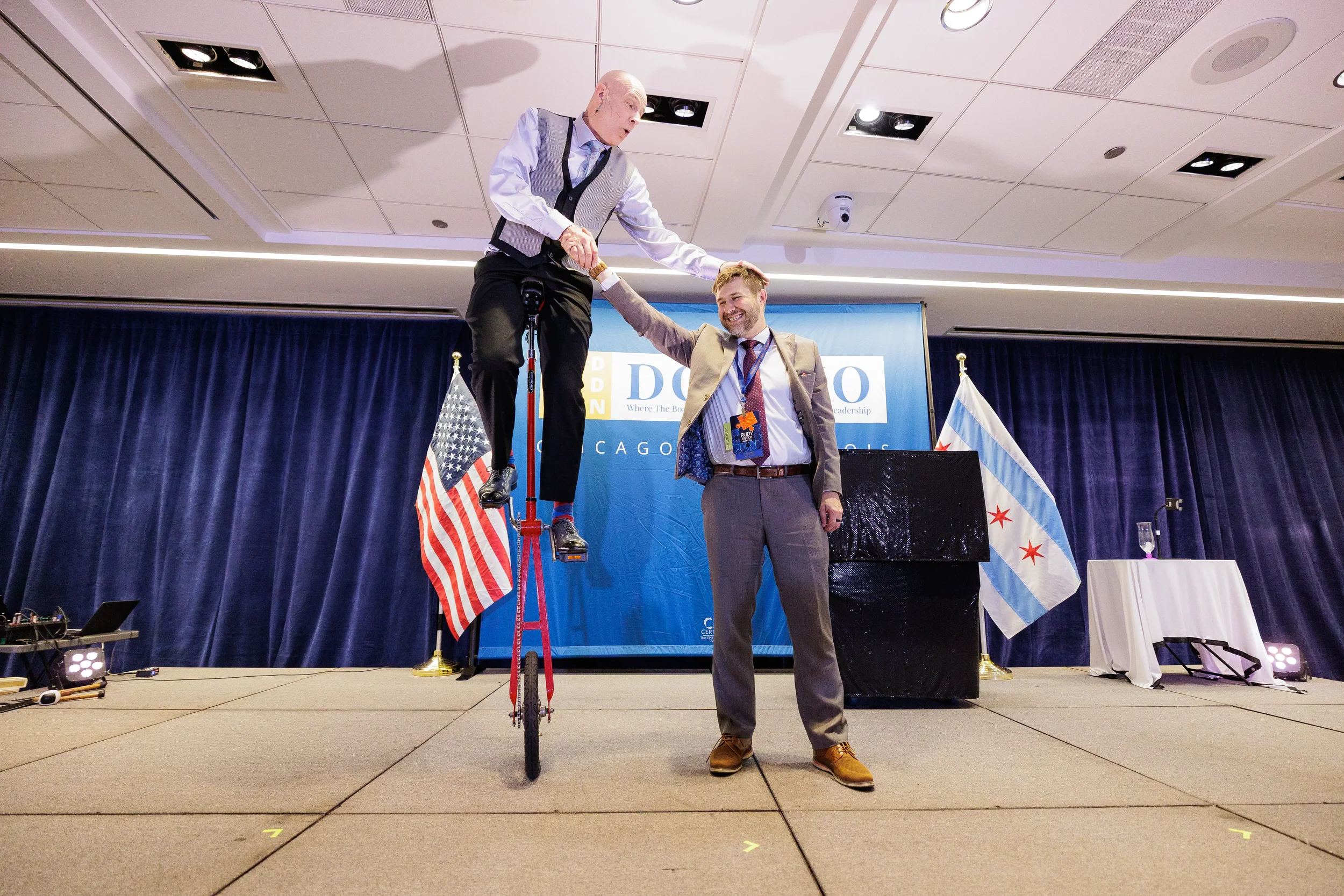 Entertainer rides unicycle on branded stage beside Chicago flag at corporate event conference