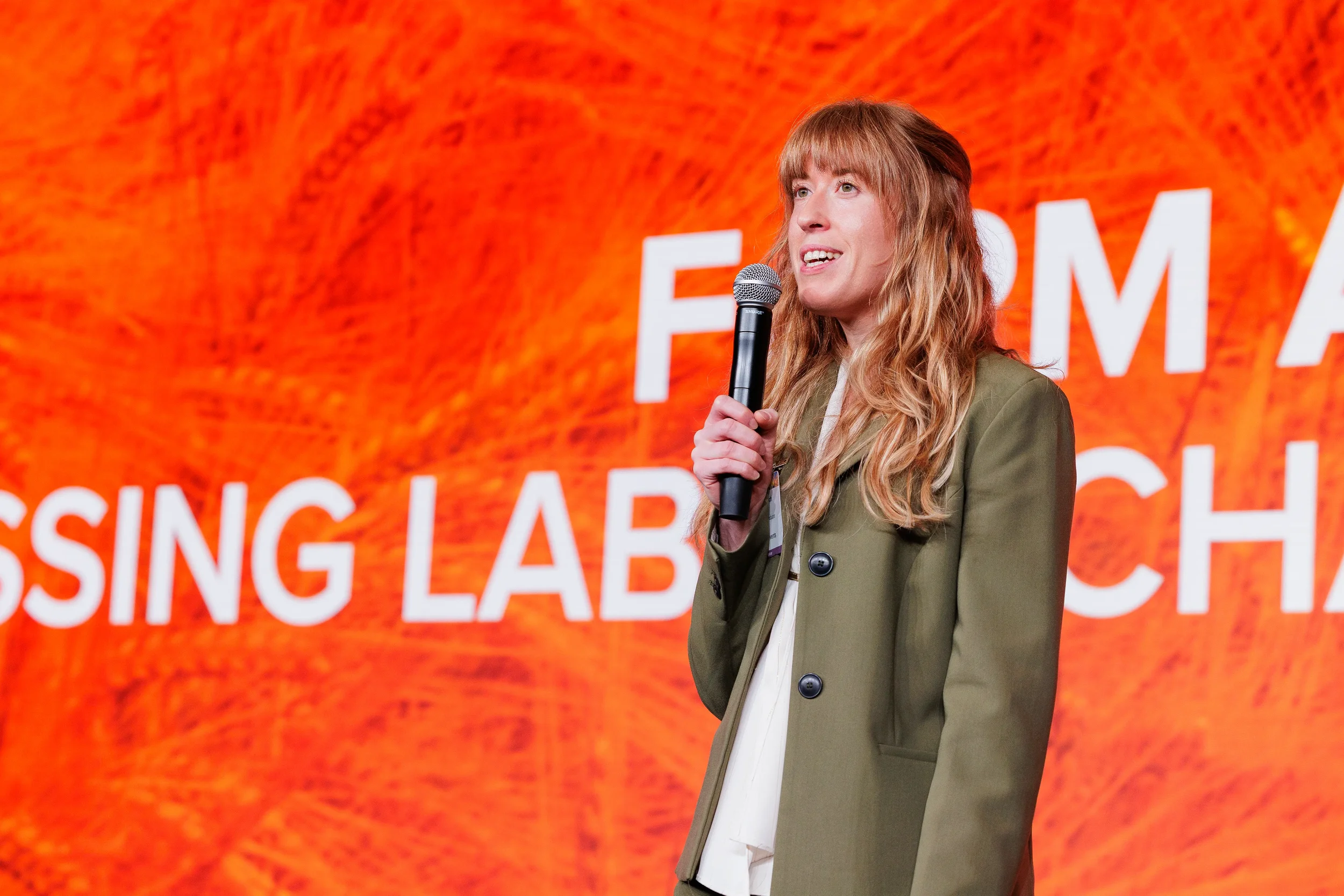 Speaker in olive blazer holds microphone on stage in front of orange branded backdrop at Orlando conference