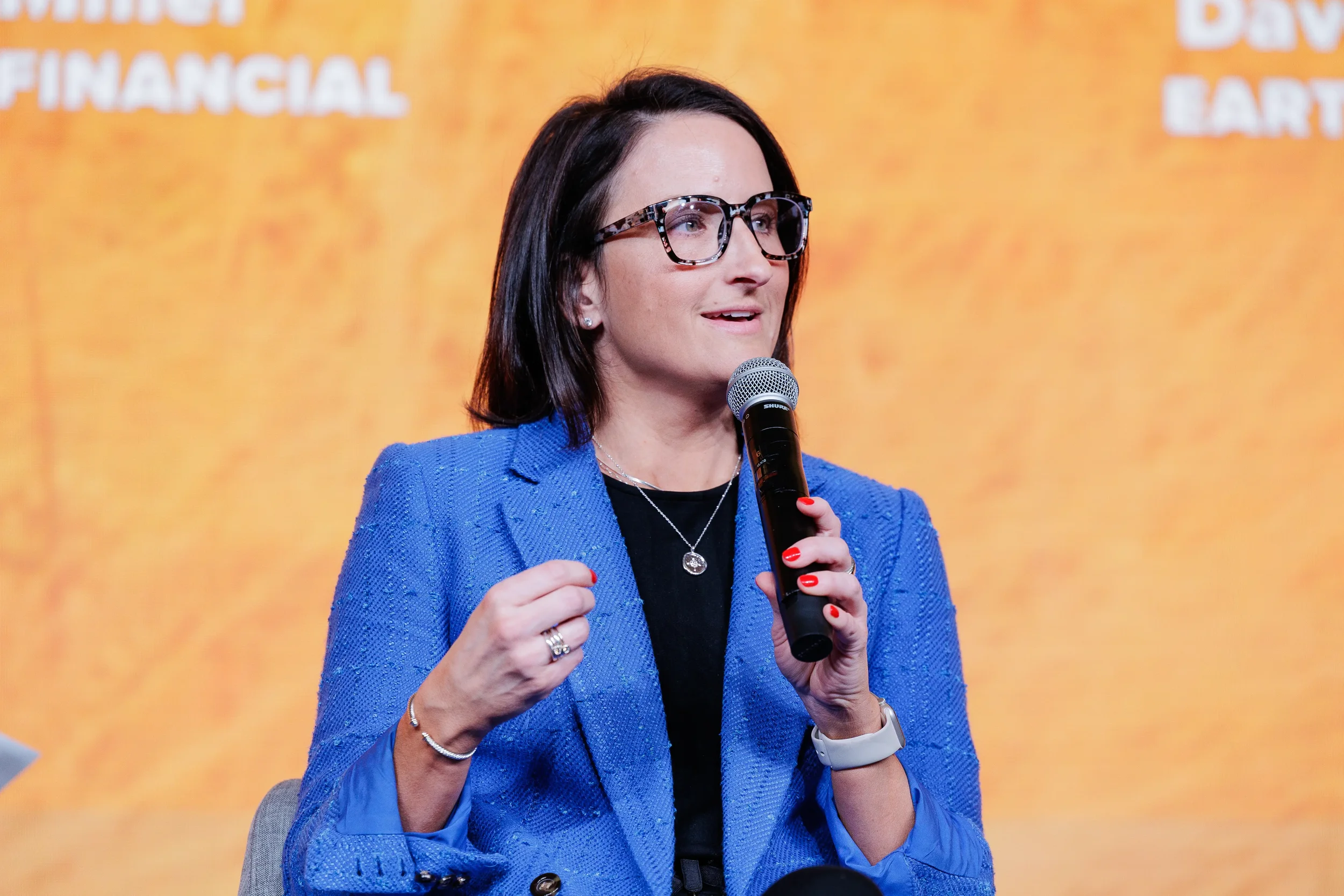 Close-up of female panelist in blue blazer holding microphone and gesturing at Orlando industry conference