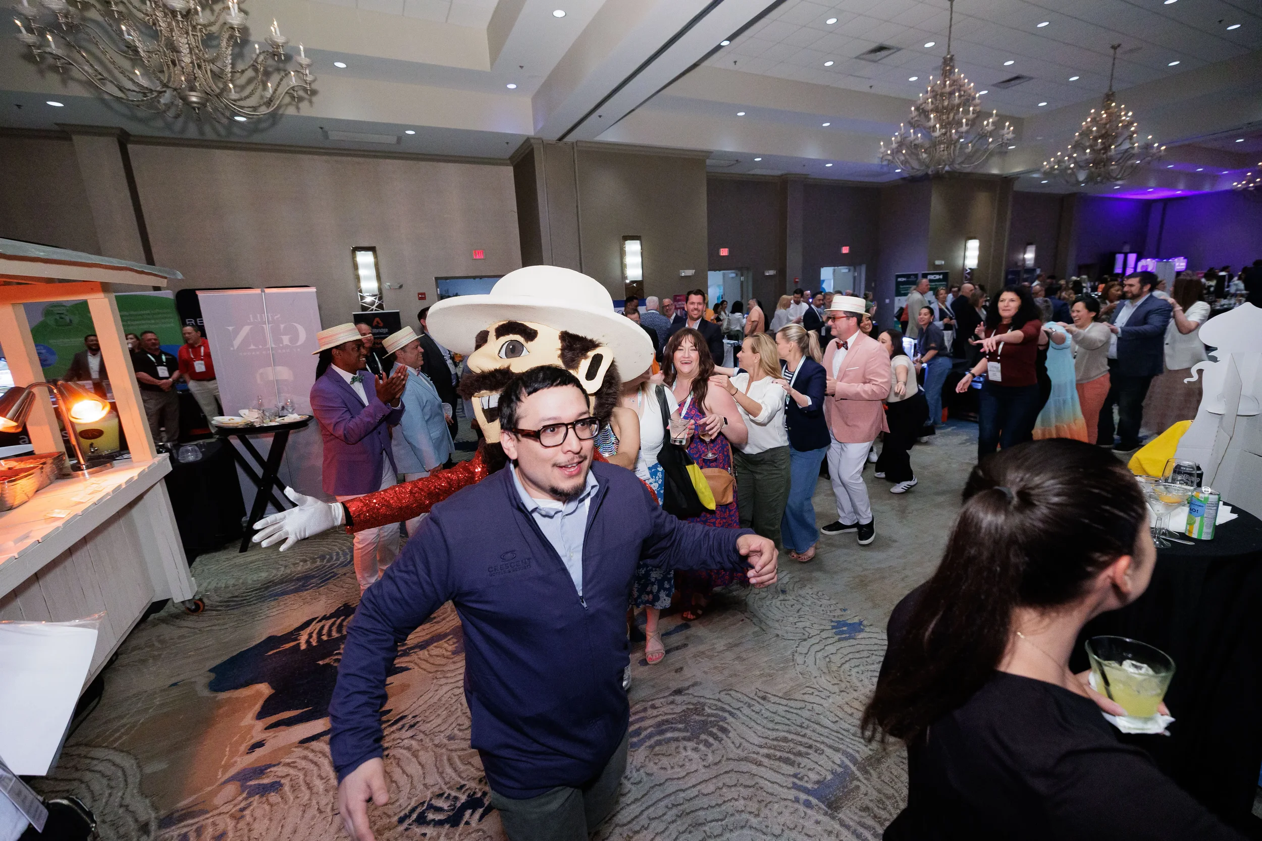 Large crowd of attendees dance alongside costumed mascot on expo floor at Orlando industry conference