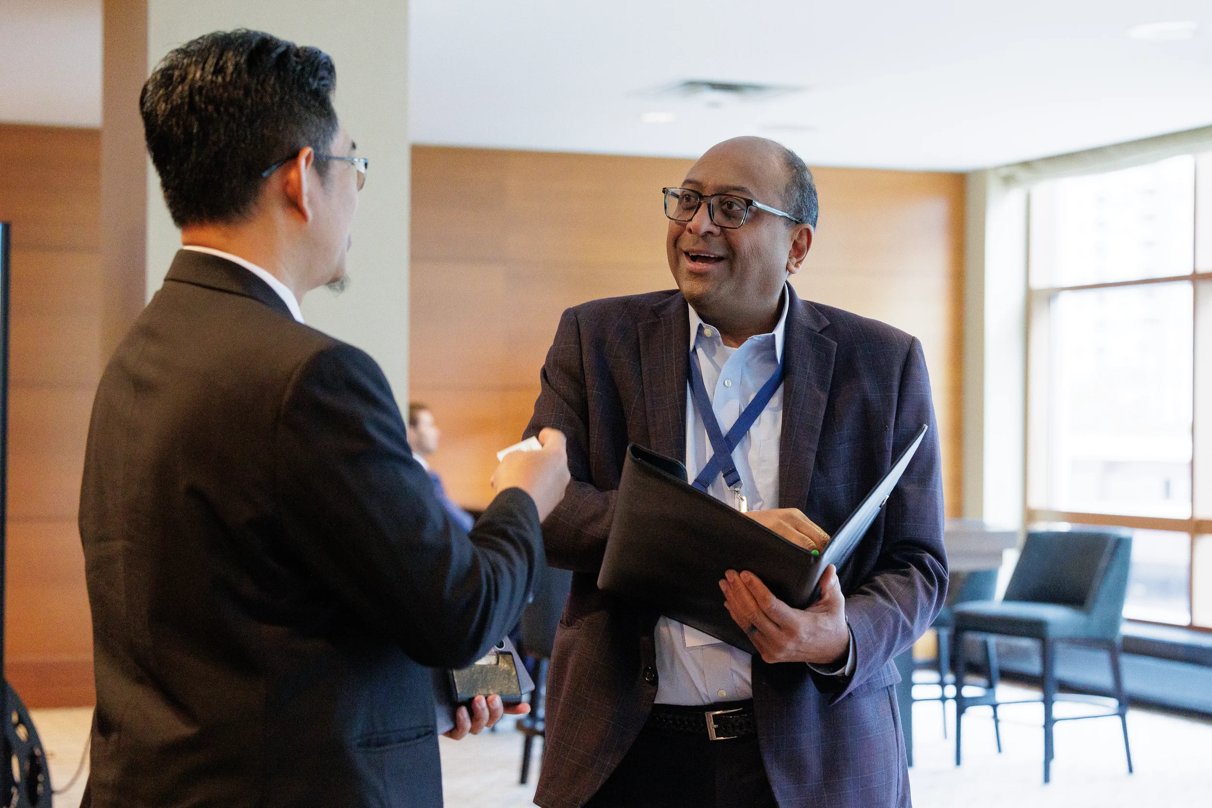 Two business men converse at a Chicago conference. One smiles while holding a folder and the other is seen from the back.
