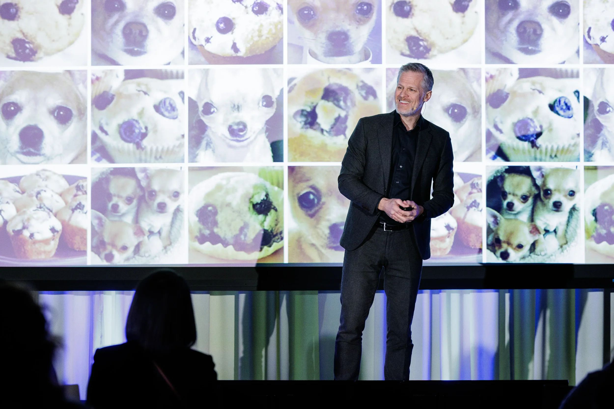 Speaker smiles on stage in front of large projection screen with audience silhouettes at Orlando event