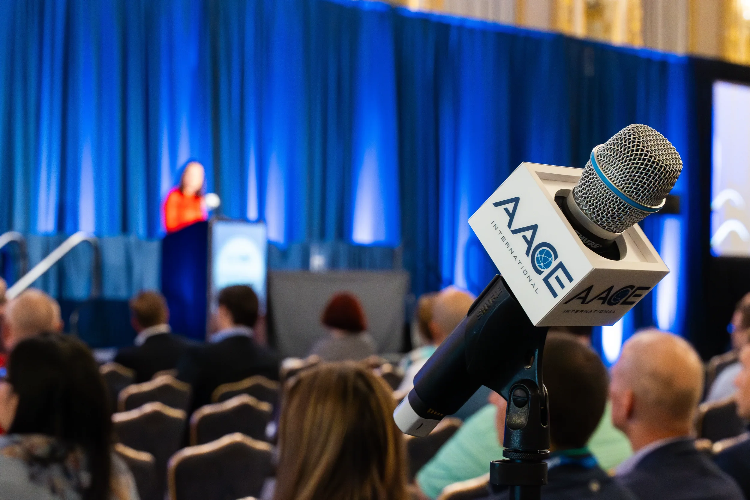 Branded conference microphone in foreground with speaker at podium and seated audience in ballroom at Orlando industry conference