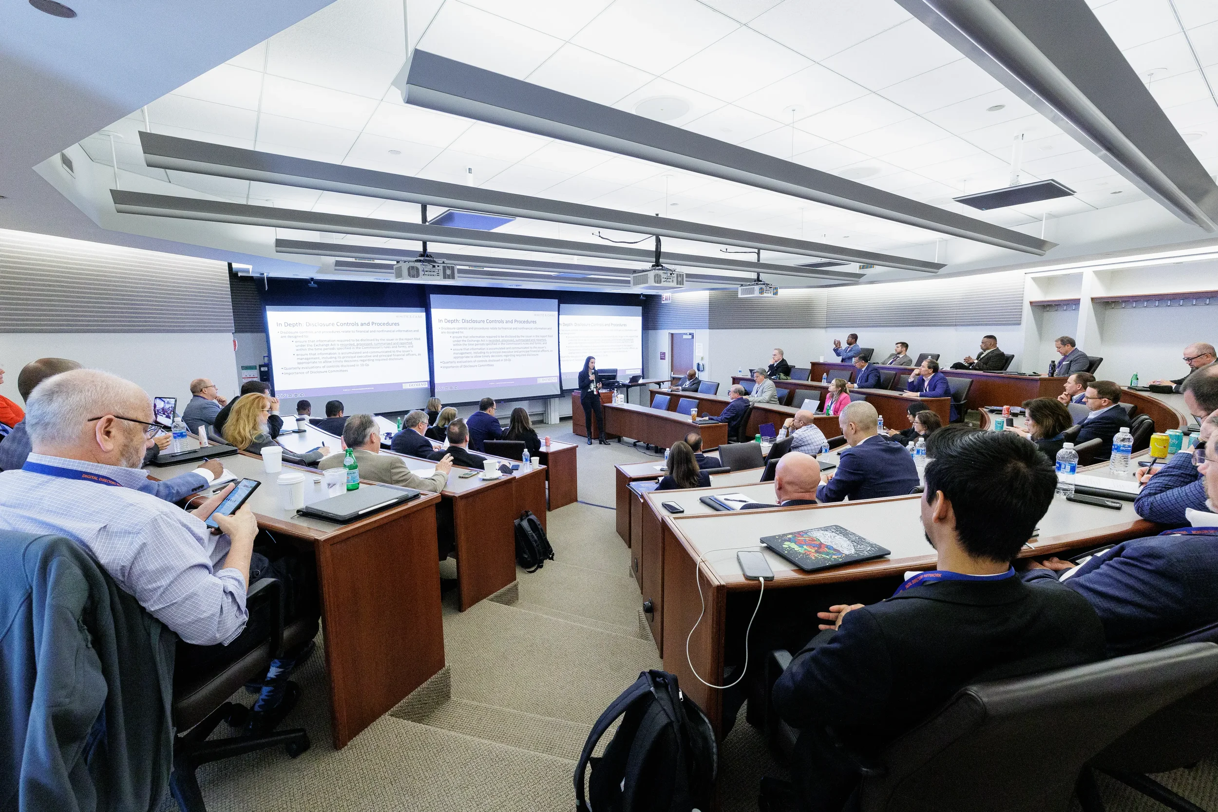 Female speaker presents to full tiered classroom with dual projection screens at Chicago corporate conference