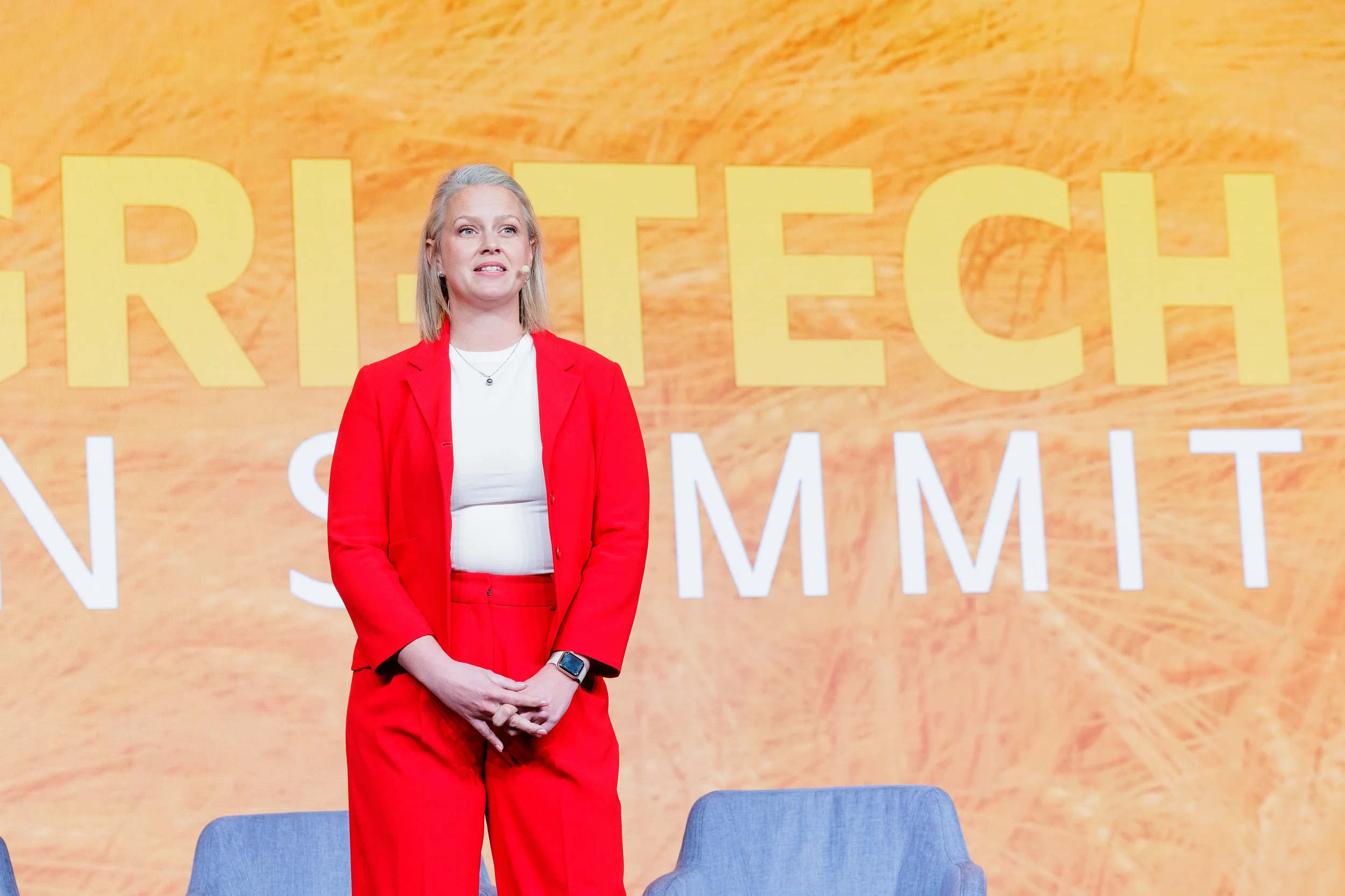 Female speaker in red suit stands before branded backdrop while presenting at Chicago industry conference