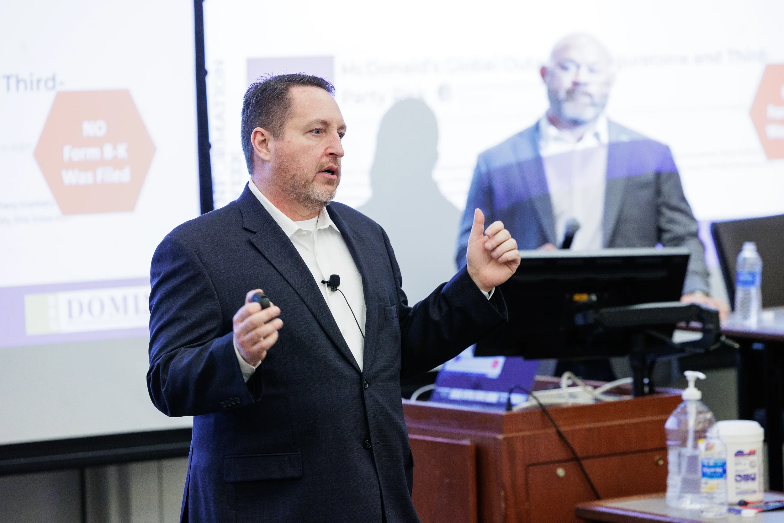 Speaker gestures with clicker beside podium during Chicago corporate conference presentation with projection screen