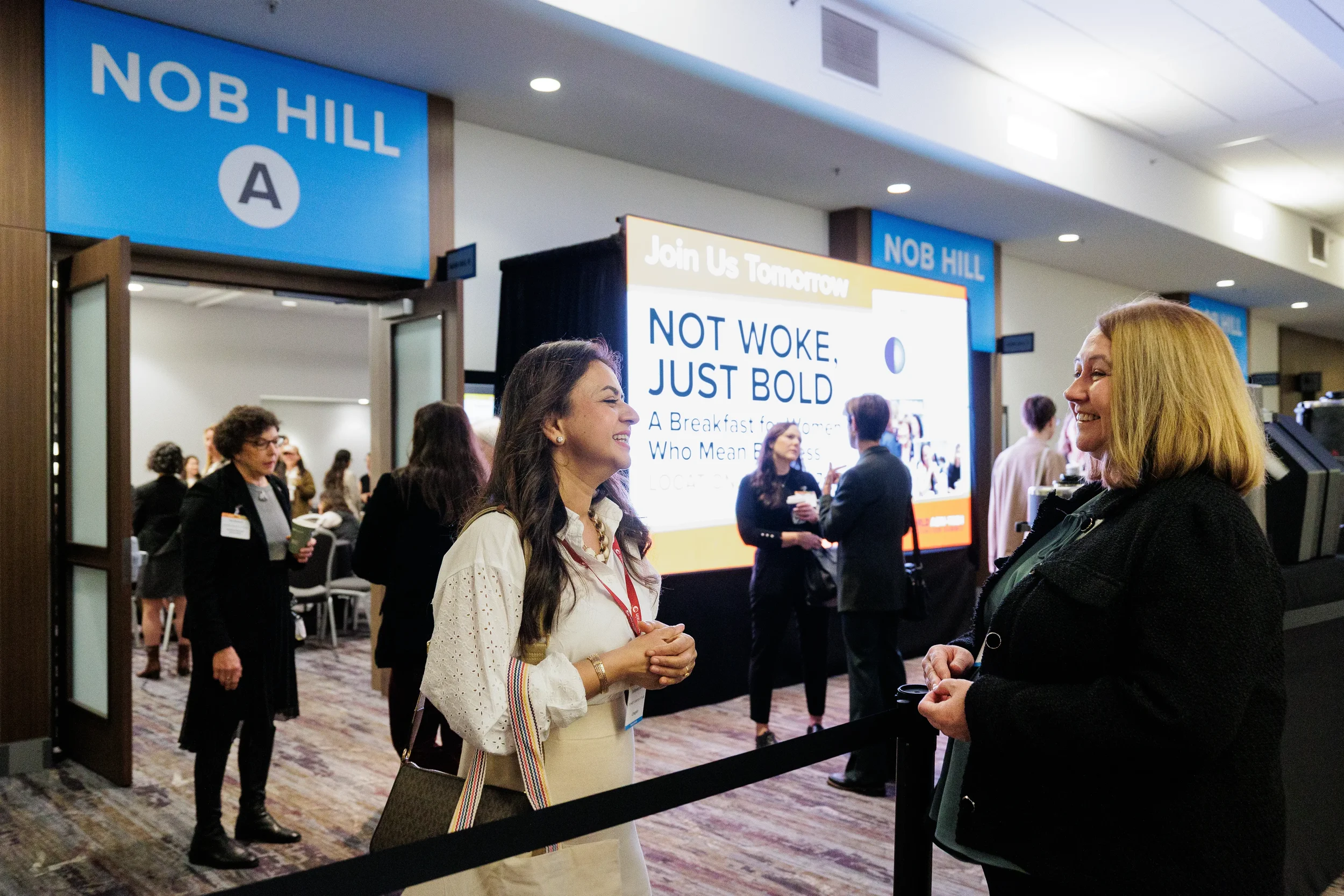 Two smiling female attendees chat near breakout room entrance and digital signage at Chicago corporate conference