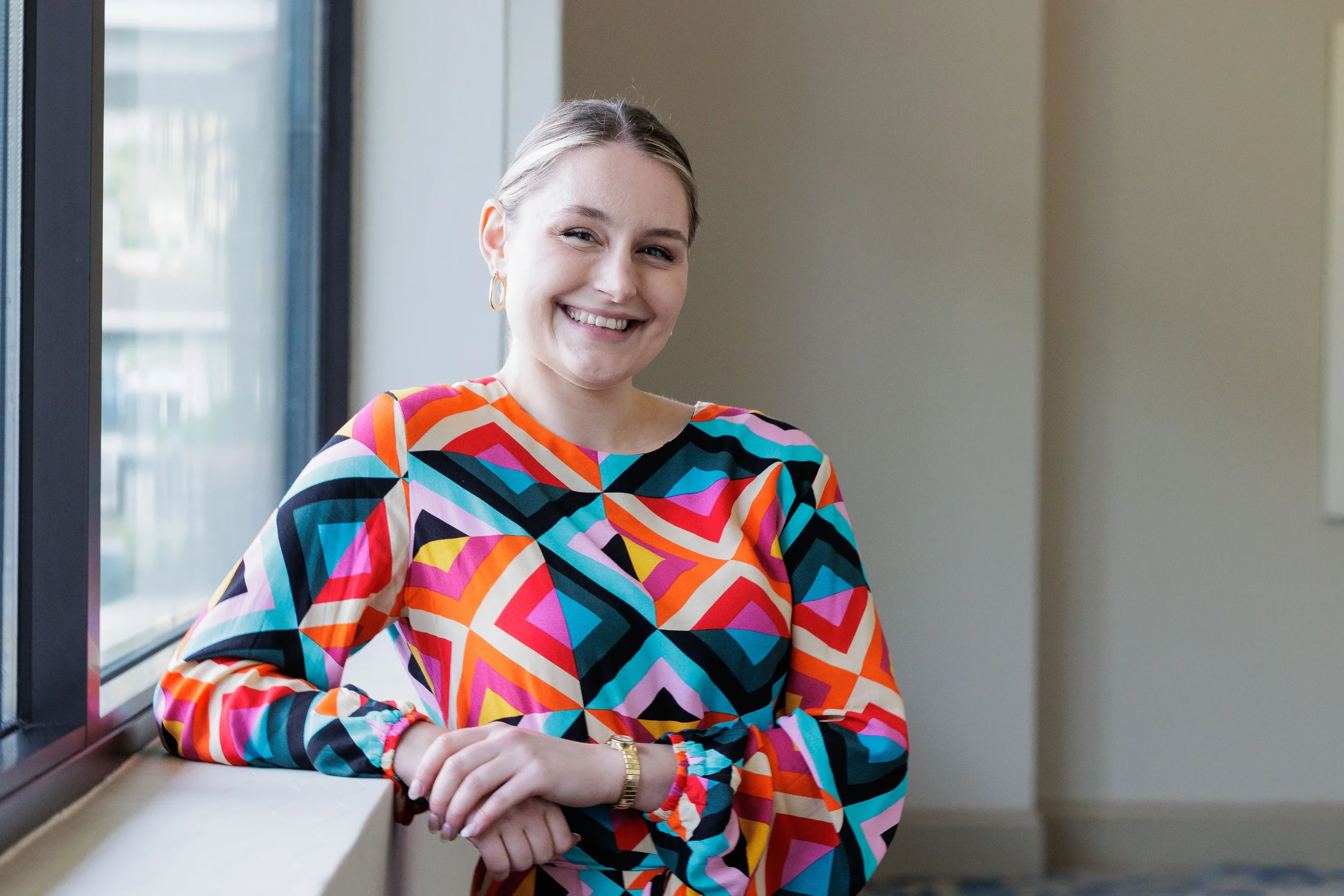 Smiling attendee in colorful geometric top leans against window at Chicago corporate conference