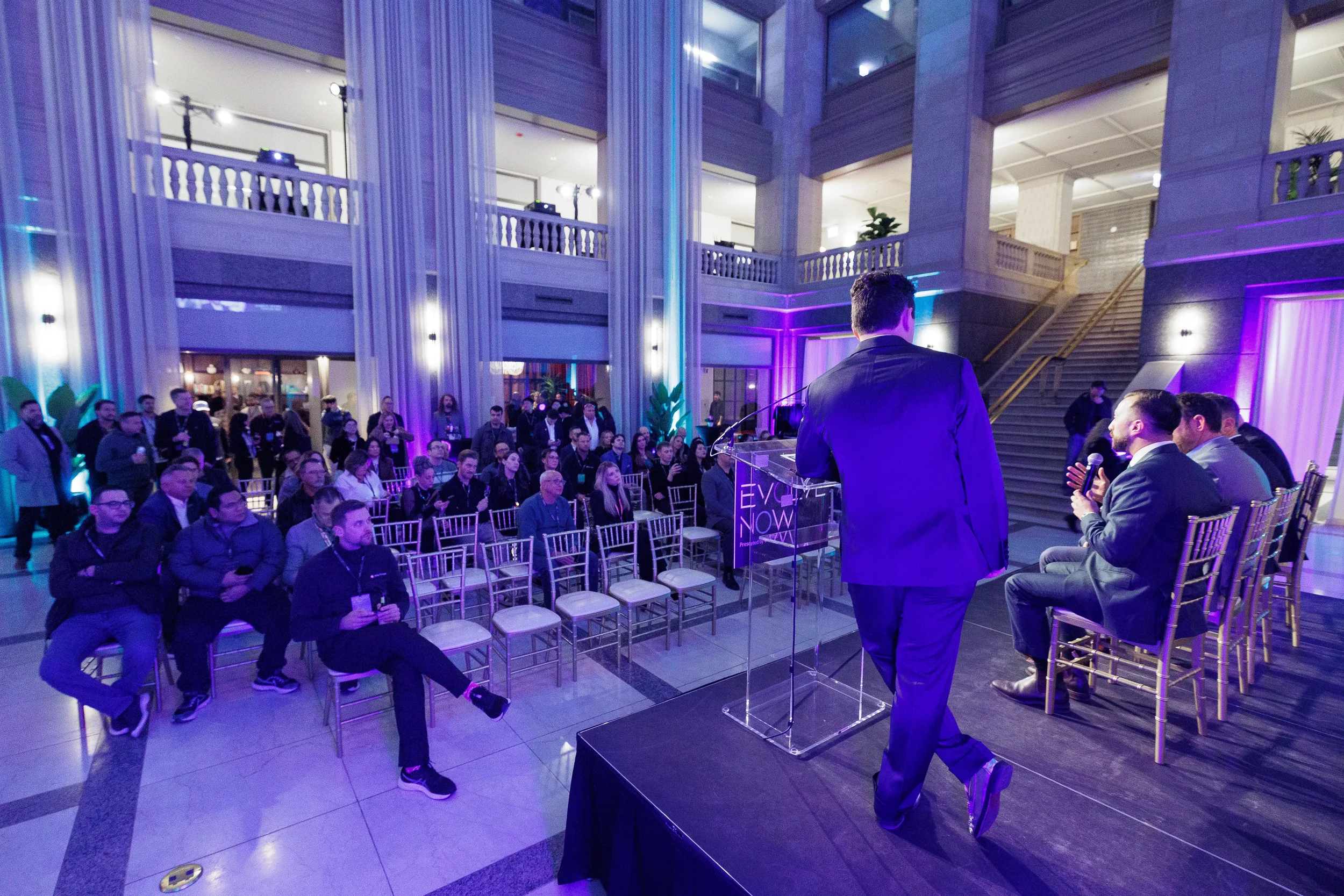 Host addresses large seated audience in grand atrium with purple uplighting at Chicago corporate event
