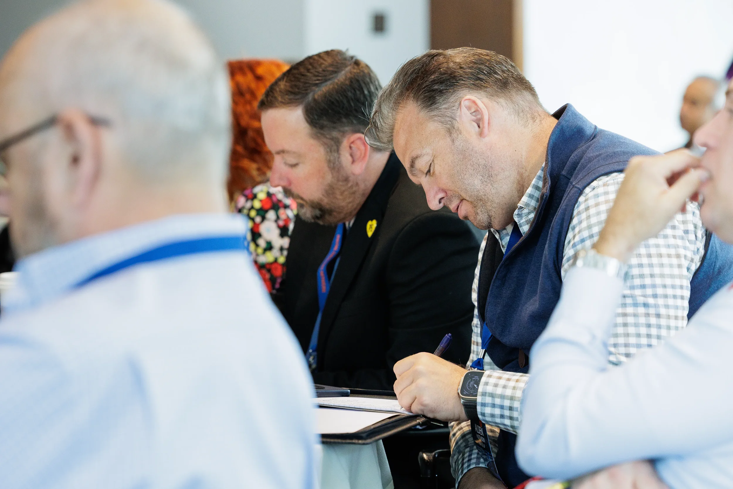 Two male attendees with lanyards write notes while seated during Orlando corporate conference session