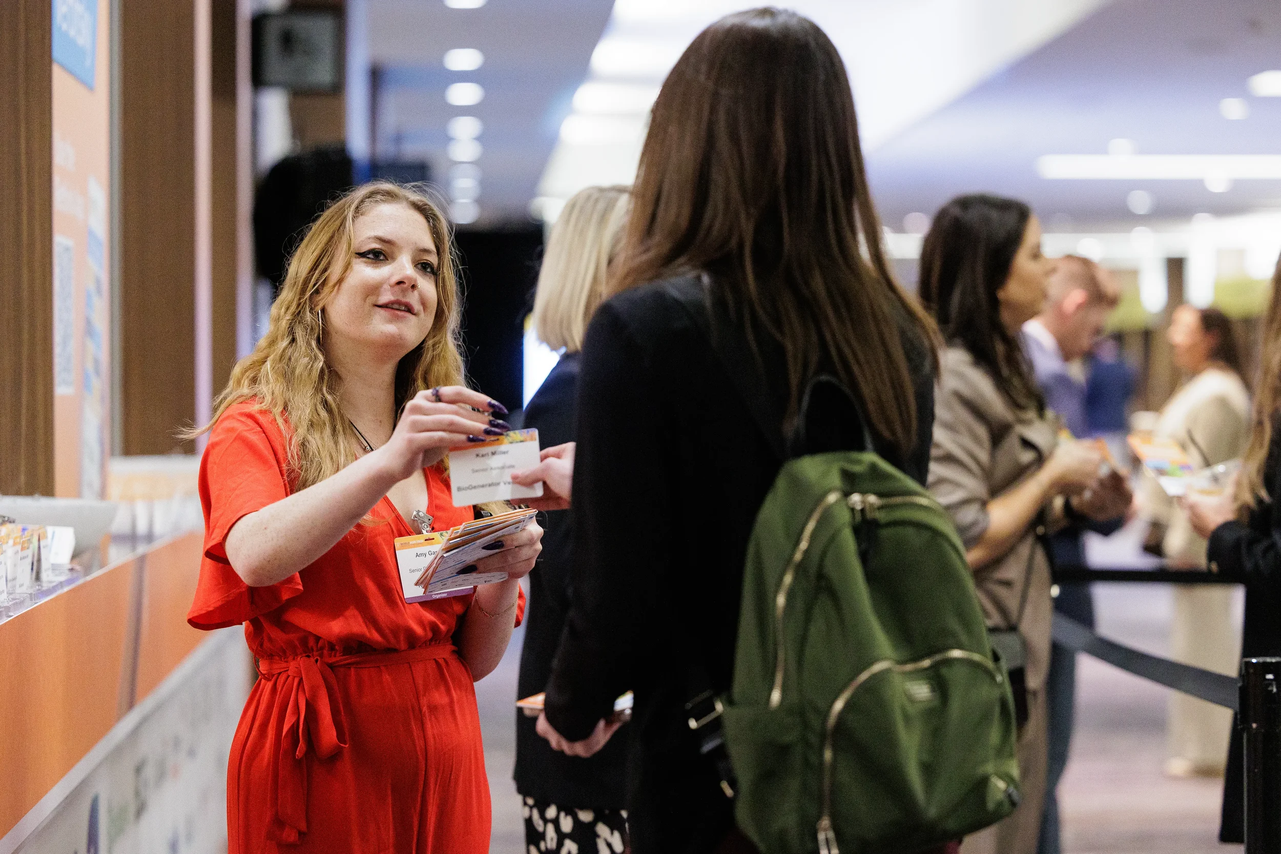Badged exhibitor in red dress shares materials with attendee in busy Orlando corporate conference hallway