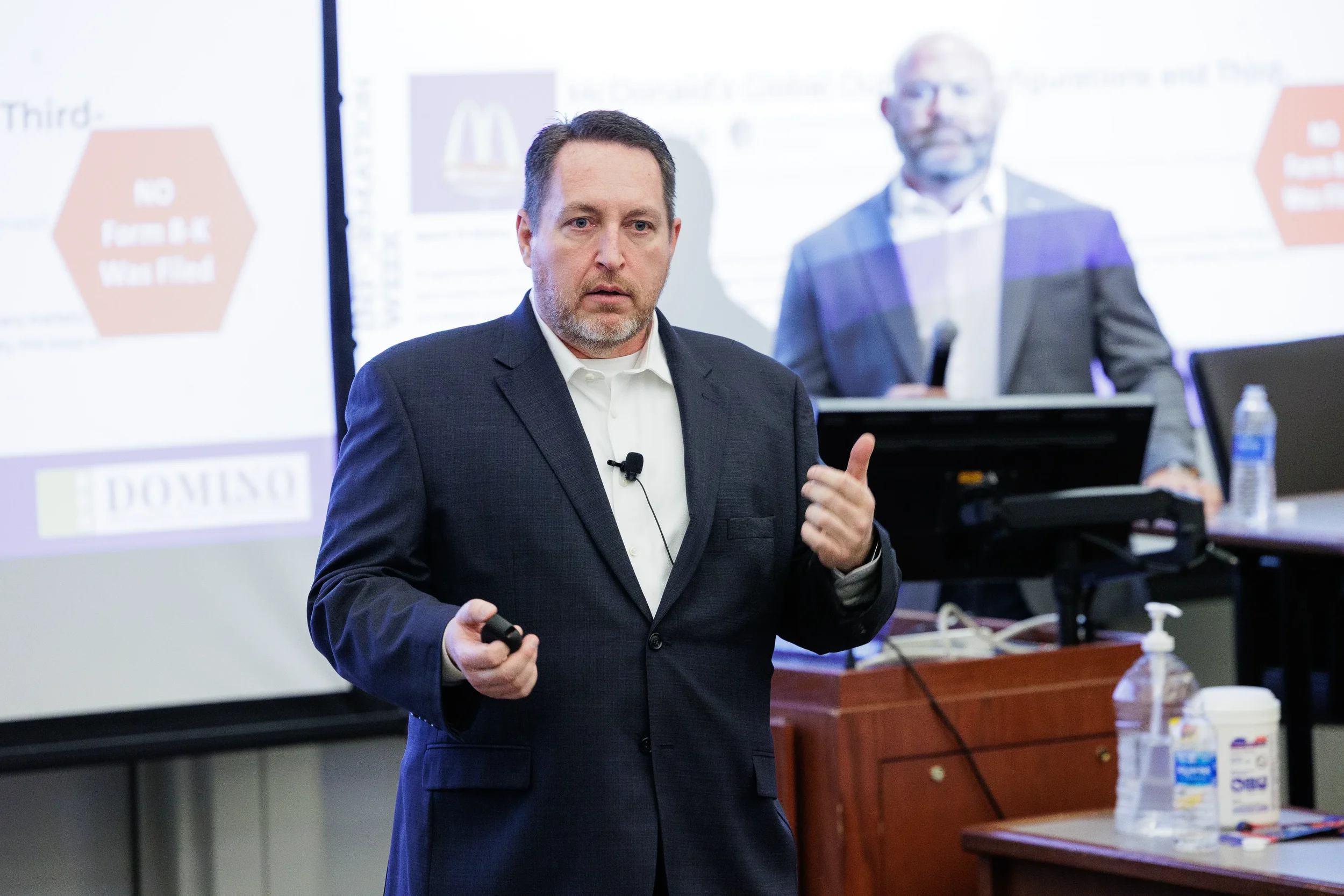Male speaker gestures with thumbs up in front of projection screen at Orlando industry conference session