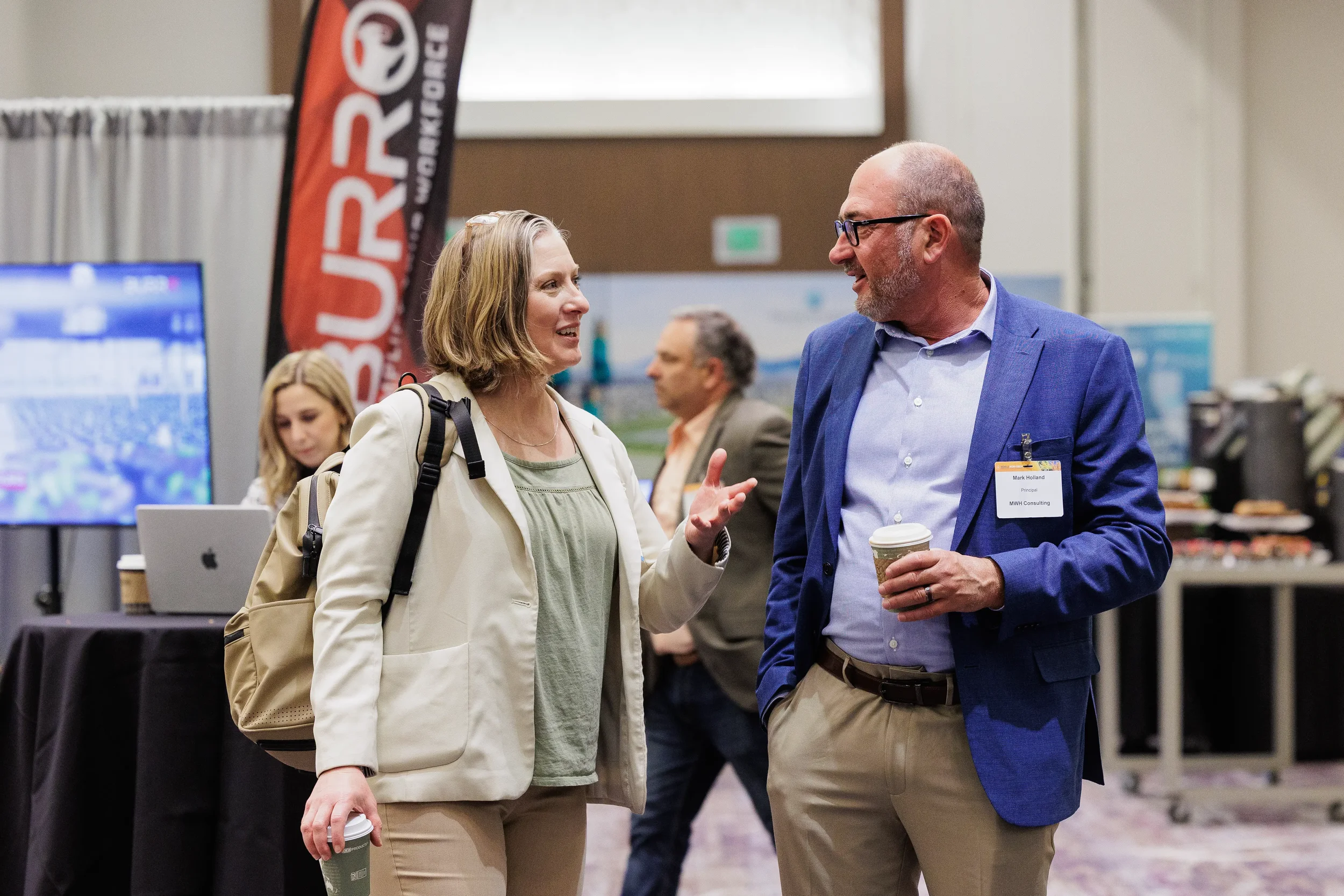 Two attendees with name badges chat over coffee on trade show expo floor at Chicago industry conference