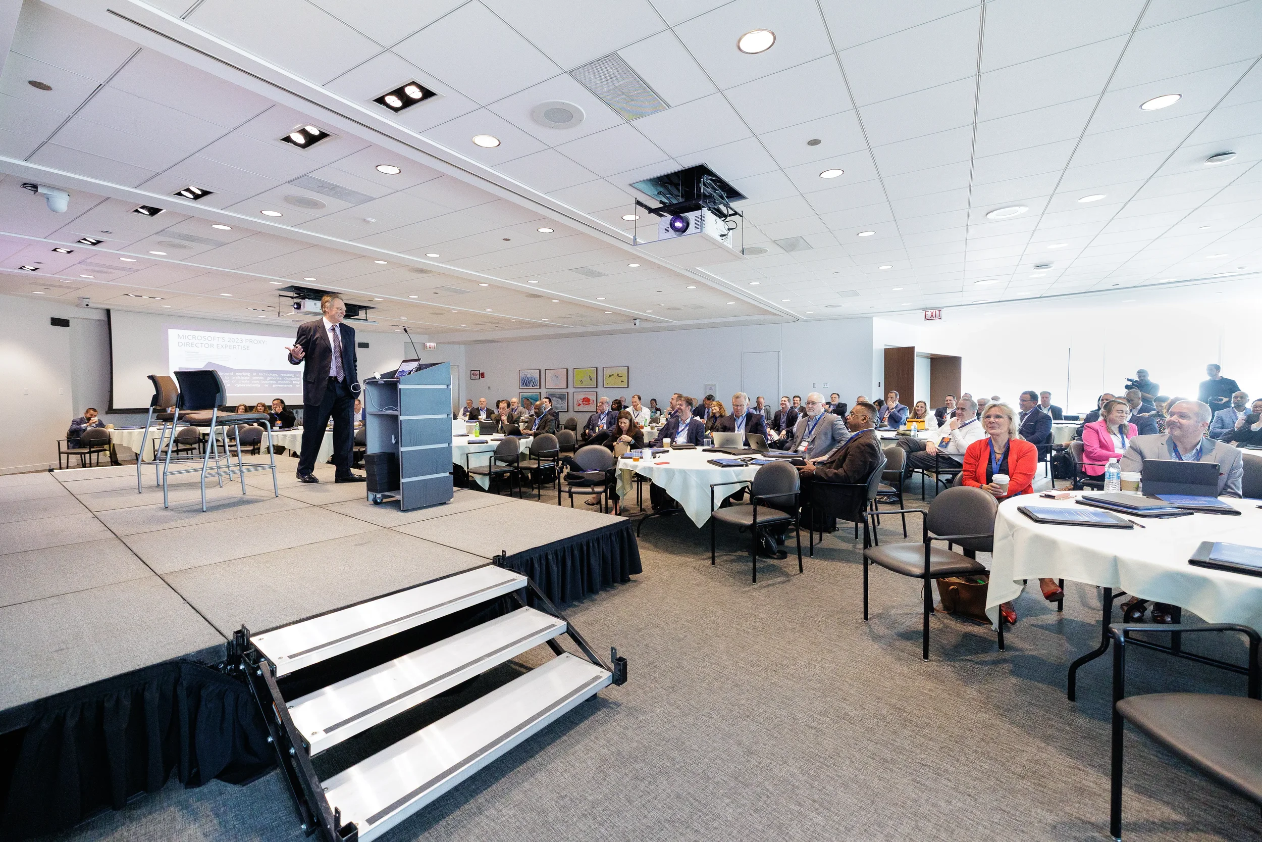 Speaker presents from elevated stage podium to full seated audience at round tables during Orlando corporate event
